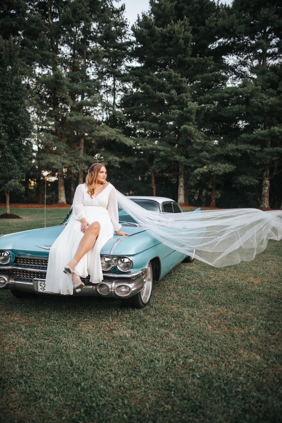 Woman in white dress sitting on vintage blue car with long veil flowing, outdoors with trees in background.