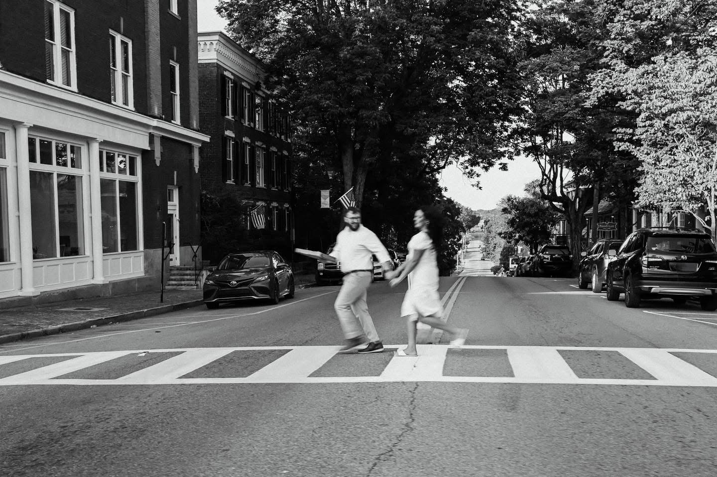Two people crossing a street on a zebra crossing, with buildings and trees in the background.