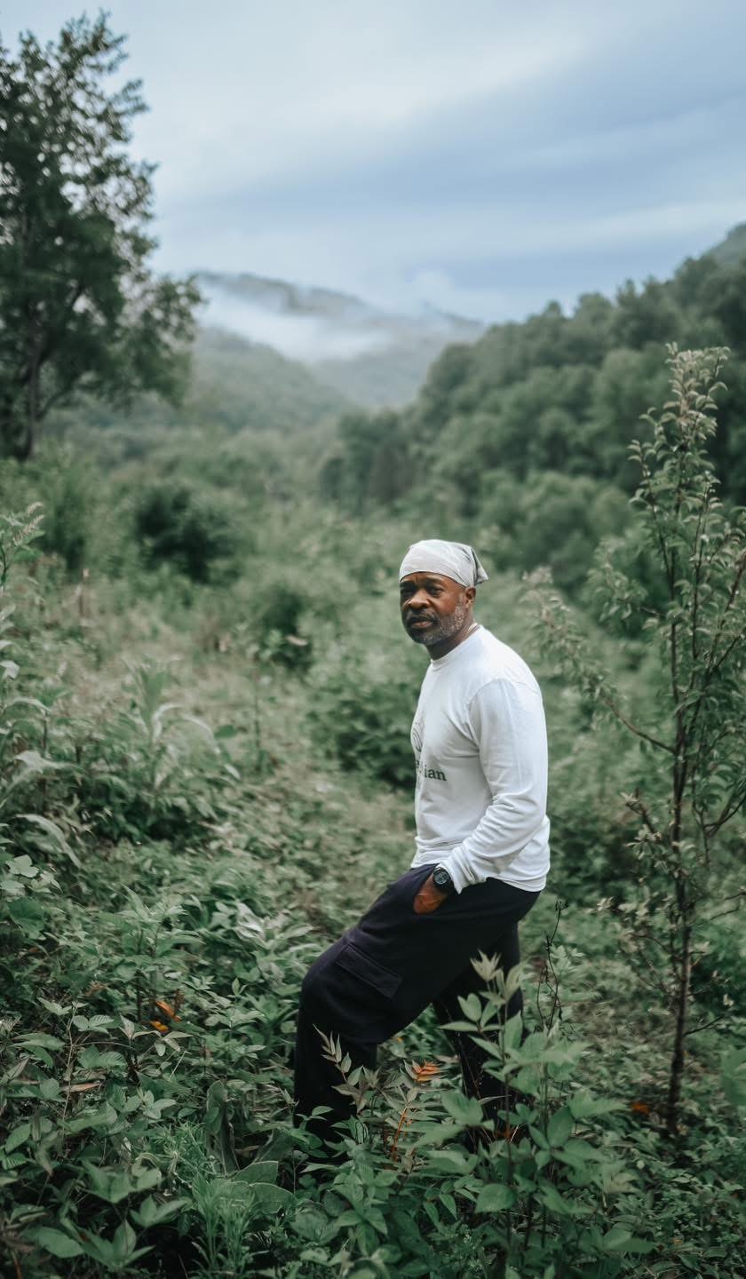 Man in white shirt and black pants standing in lush green forest with hills and mist in background.