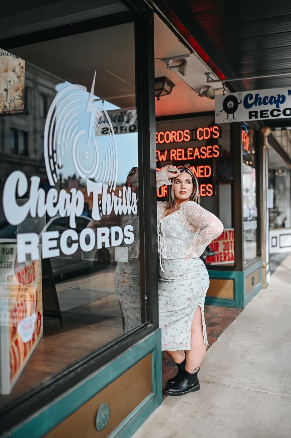 Woman standing outside a record store with large window display, sign reading 'Cheap Records,' and red and black signage inside.