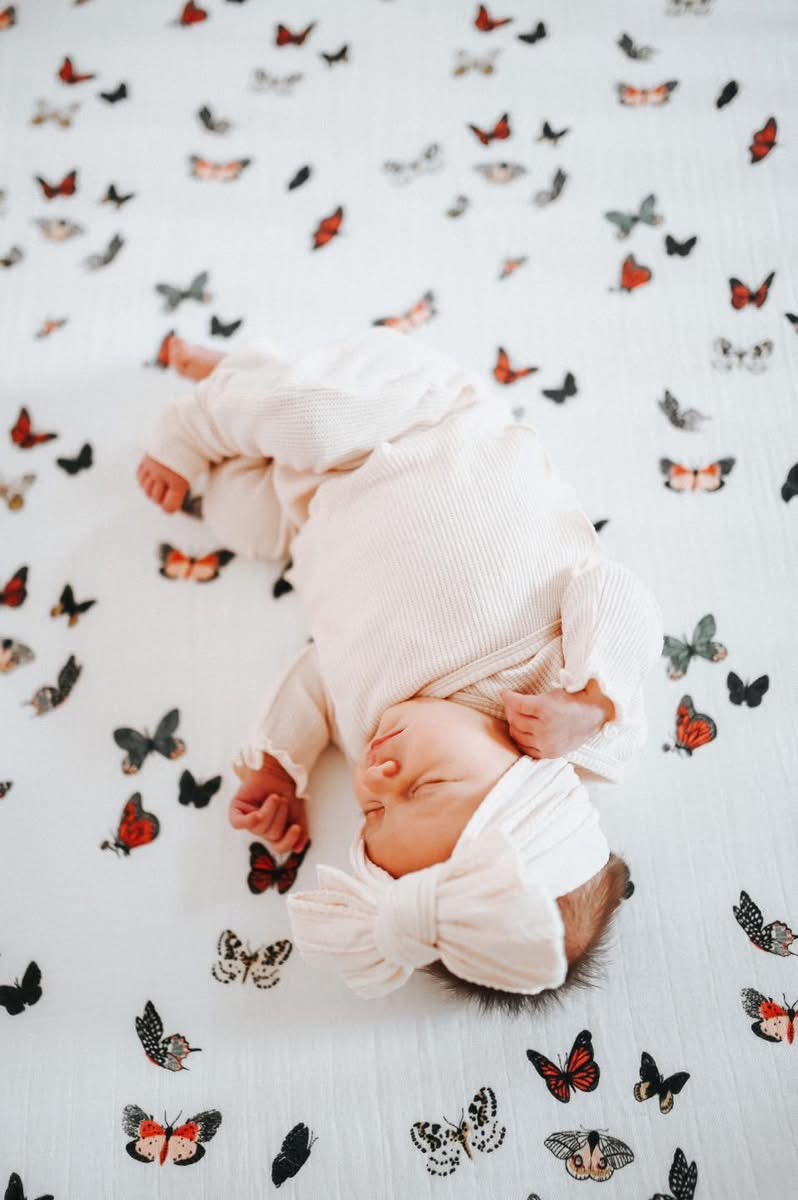 Baby lying on a butterfly-patterned blanket, wearing a white outfit and hat, sleeping peacefully.