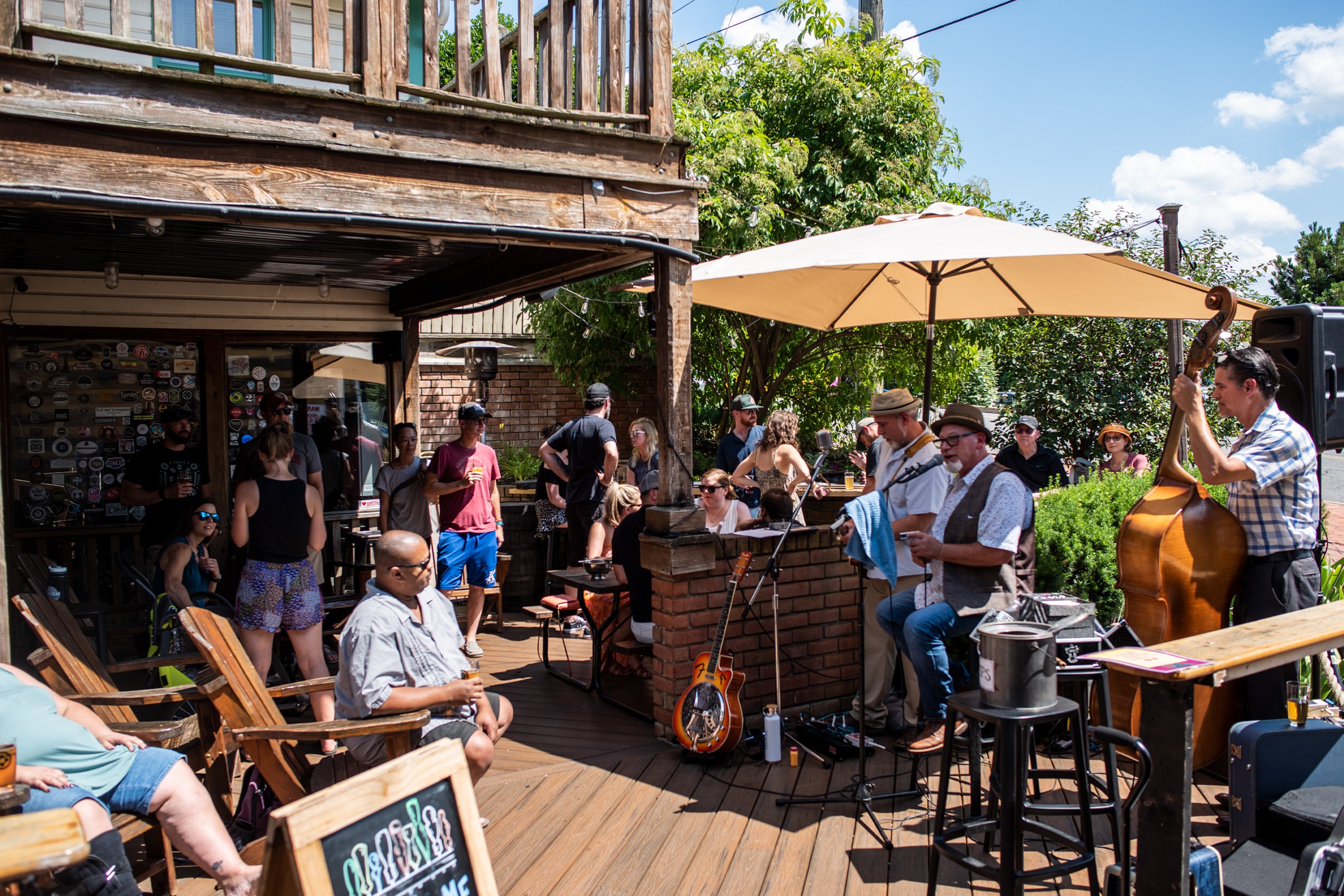 People gather outdoors at a social event with music, seating, and a large umbrella, under a wooden structure and trees.