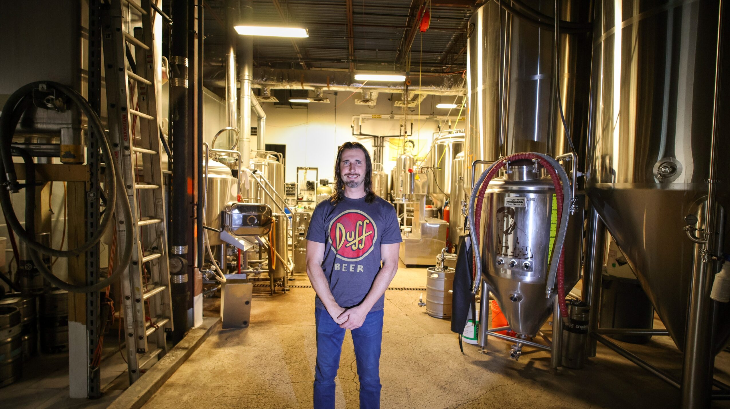 Person standing in a brewery with large metal tanks and equipment around them.