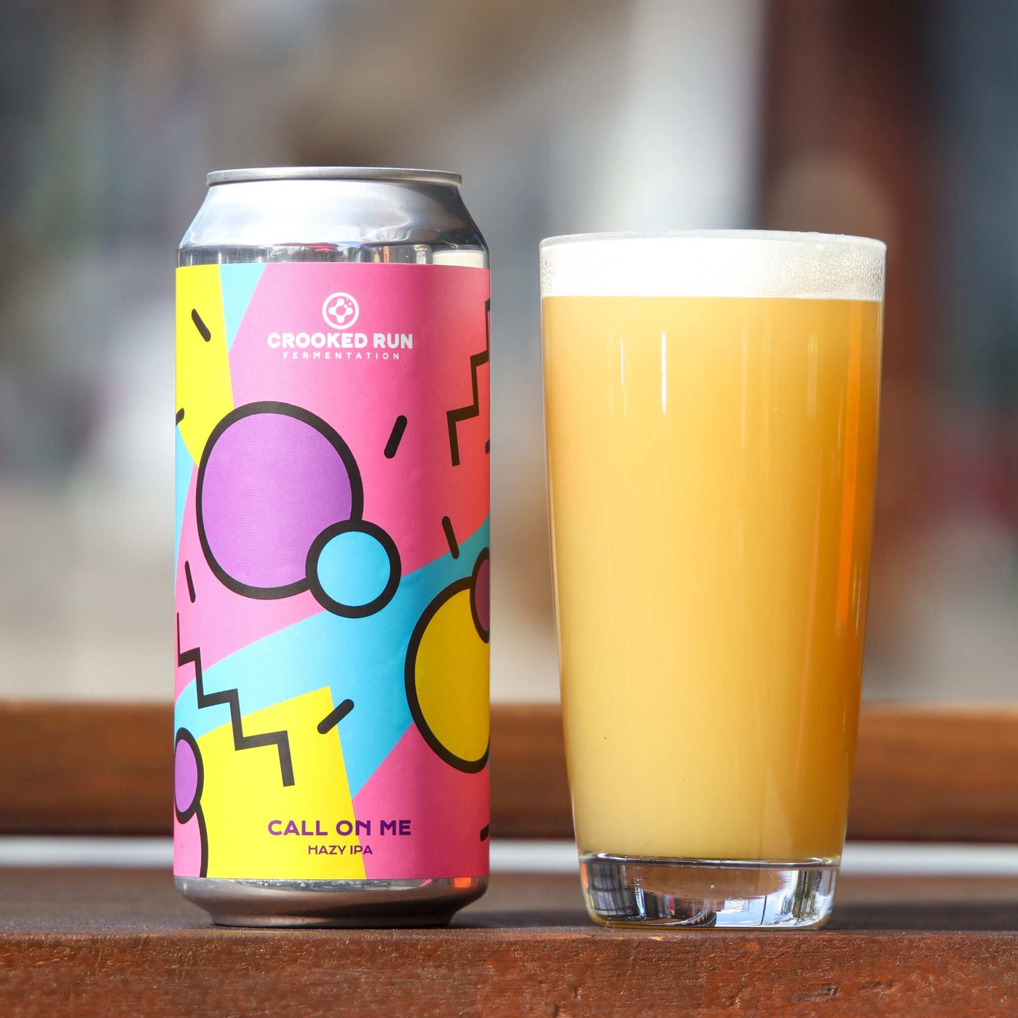 A colorful can of craft beer and a glass filled with hazy yellow beer on a wooden surface.