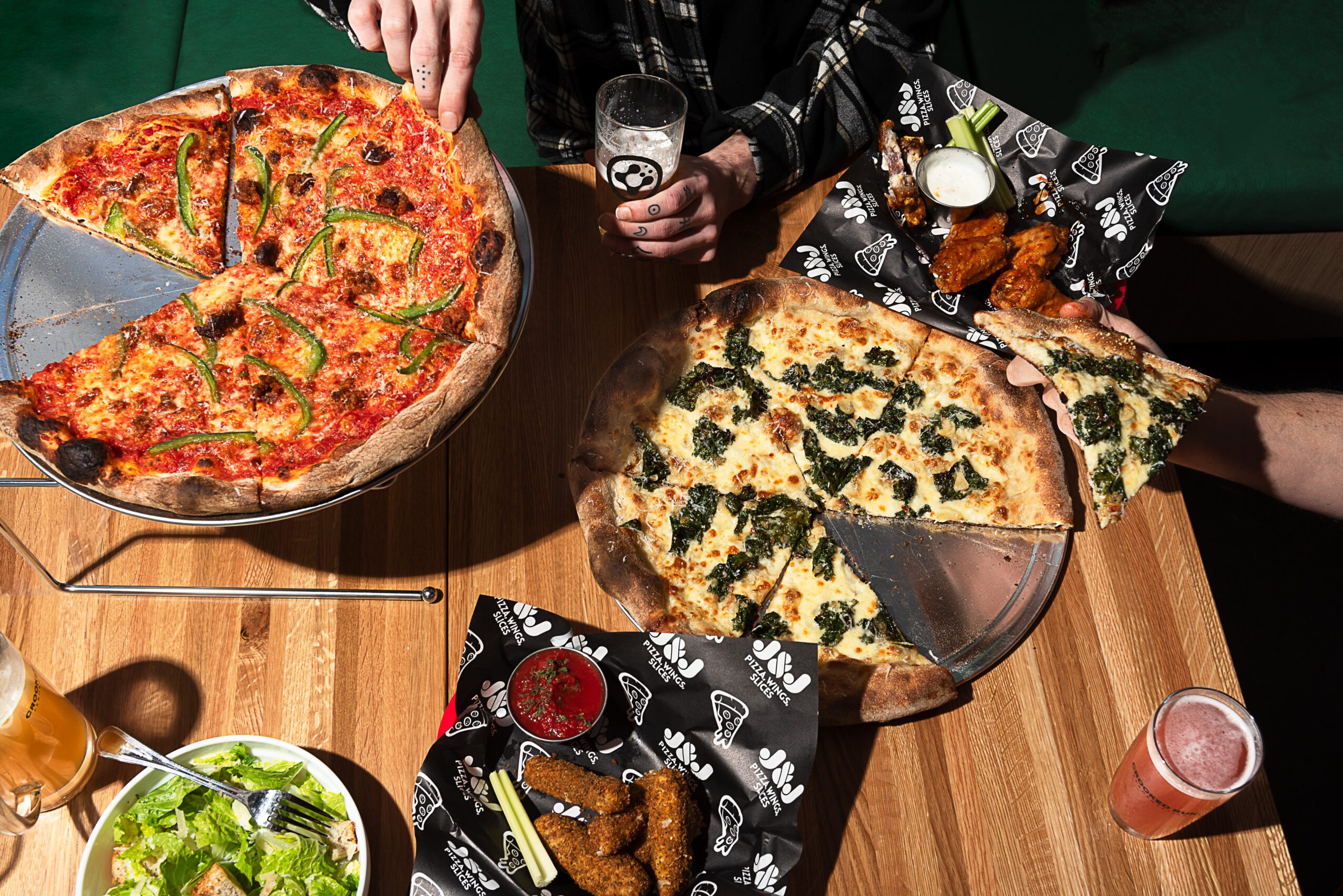 Two people share a table with pizza, fried chicken, and drinks, with hands reaching for food and drinks on a wooden table.