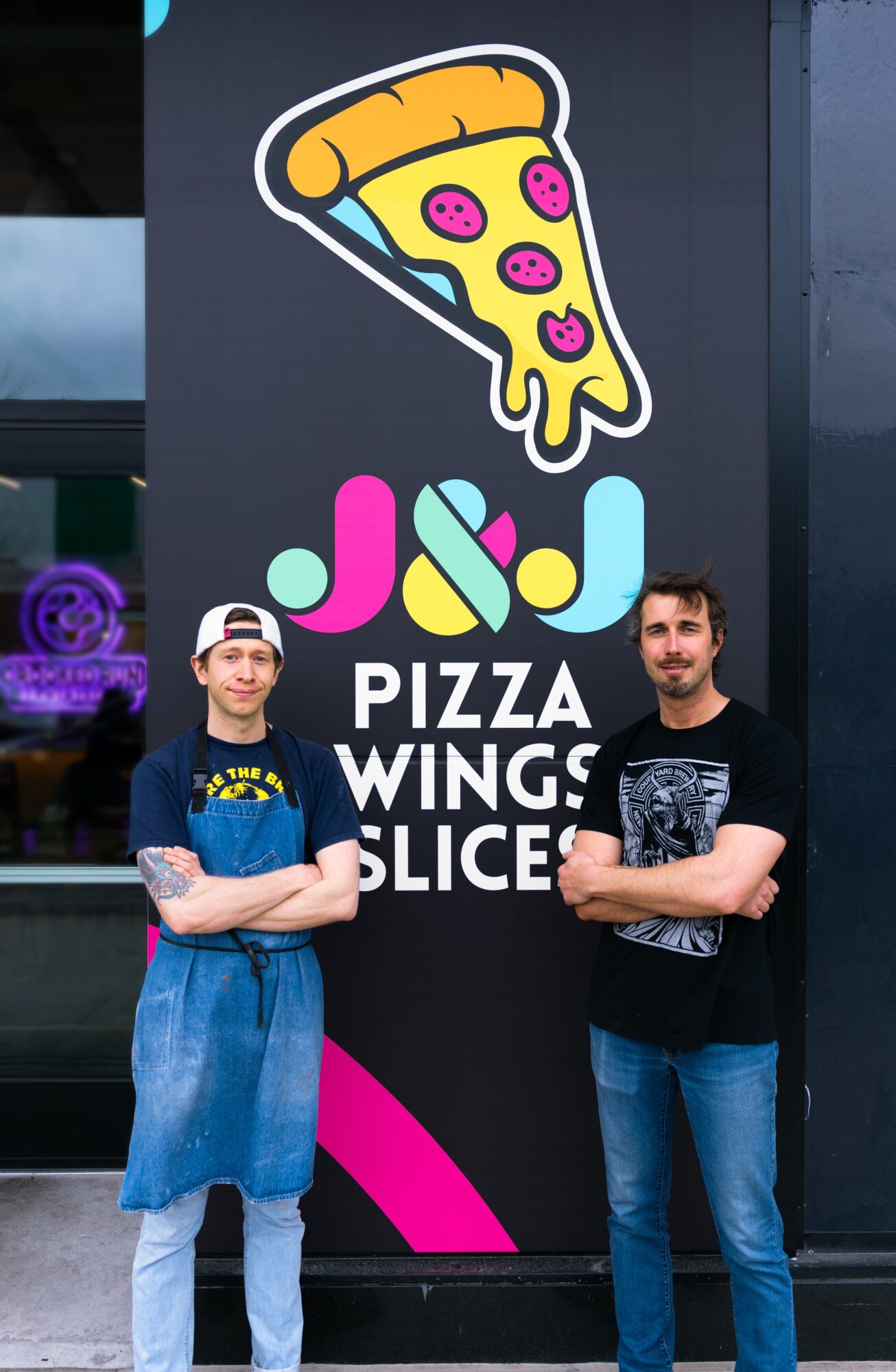 Two men stand in front of a pizza restaurant sign with a pizza slice and colorful logo.