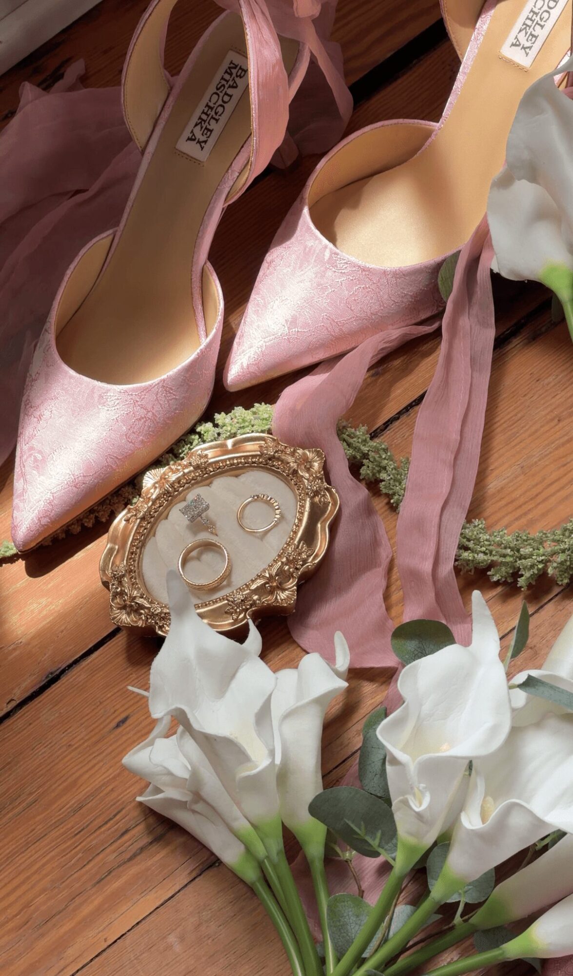 Pink high-heeled shoes with ribbons, a decorative mirror, and white flowers on a wooden surface.