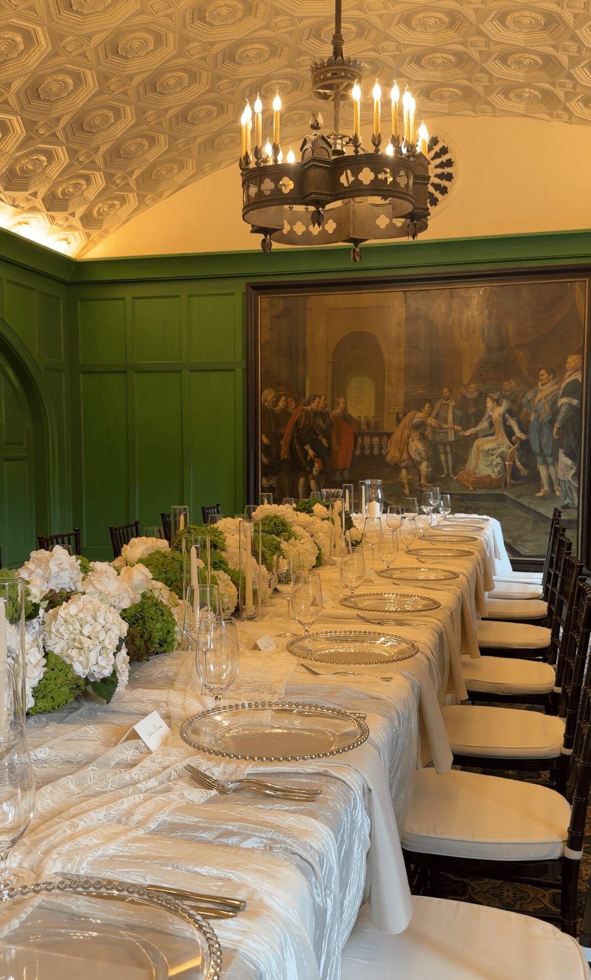 Long dining table with white tablecloth, floral centerpieces, and chairs in a decorated room with green walls and a chandelier.