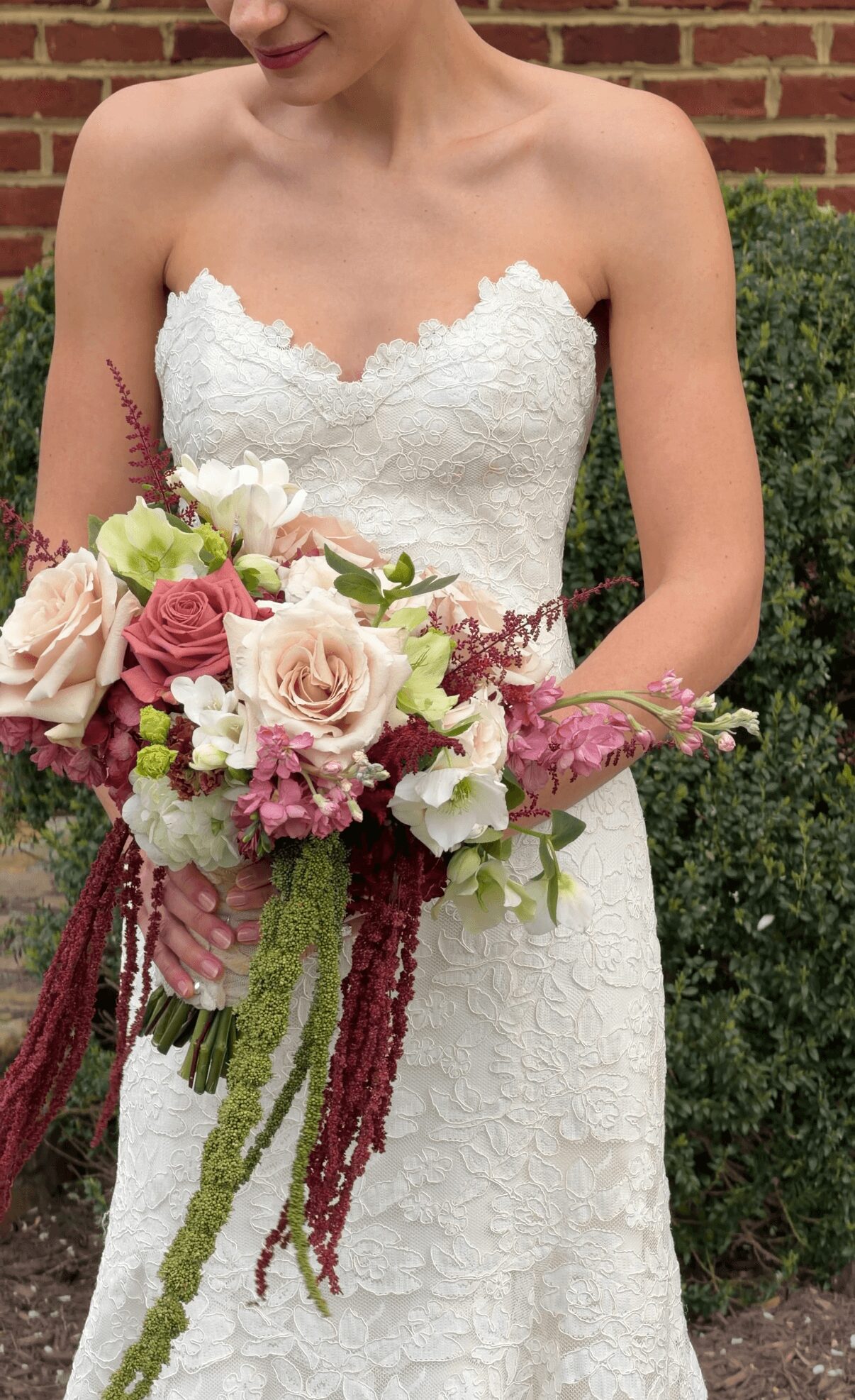 Bride in a strapless lace wedding dress holding a bouquet of roses, peonies, and greenery outdoors.