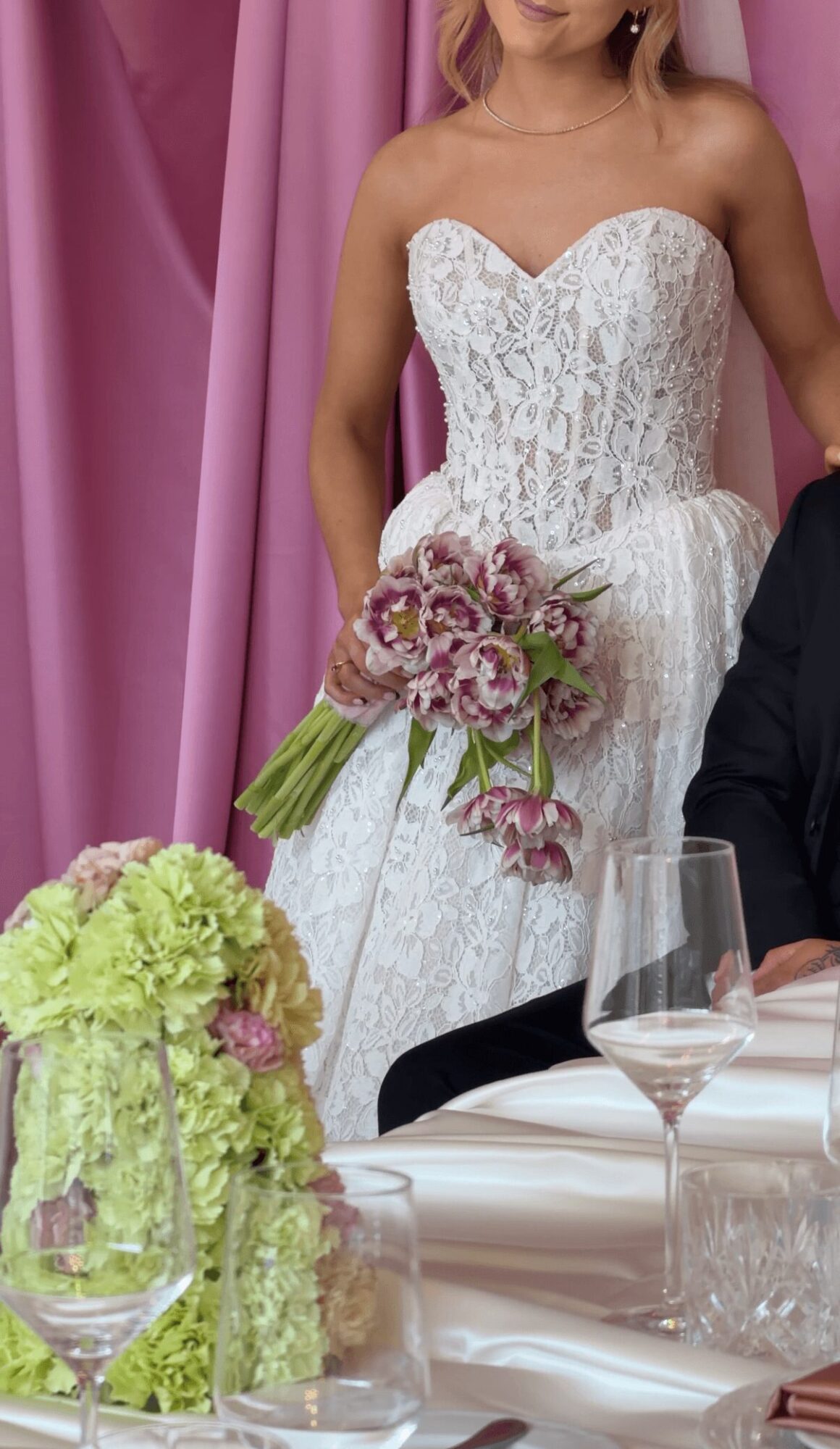 Bride in a white lace wedding dress holding a bouquet, standing near a table with floral arrangements and glassware.