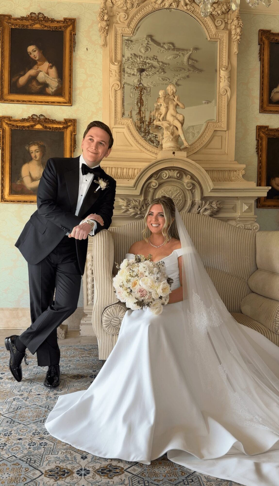 Bride seated on sofa holding bouquet, groom standing beside her, elegant room with paintings and ornate decor in background.