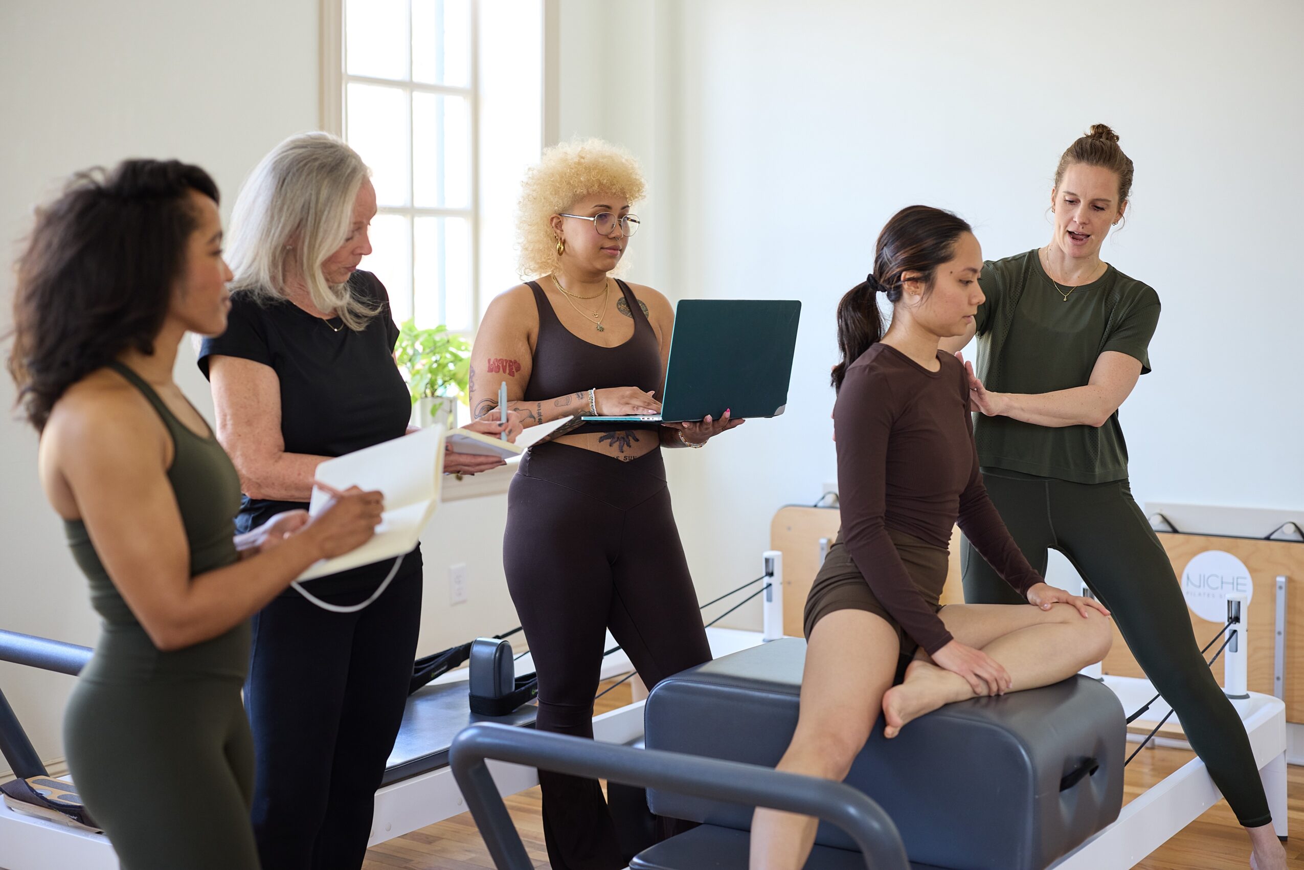 Group of five women in a medical or therapy setting, with one woman demonstrating a technique on a patient lying on a table.