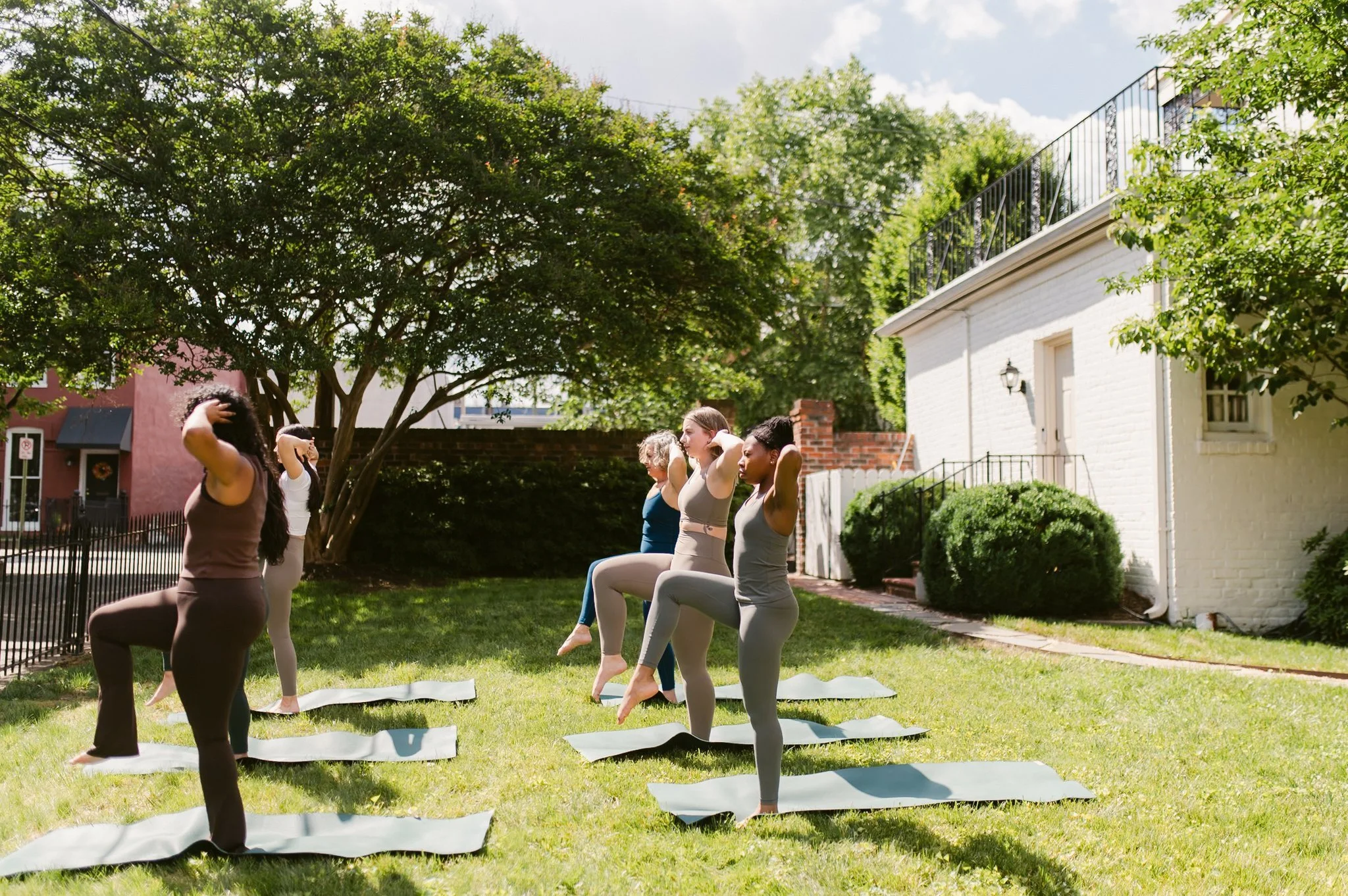 Group of people practicing yoga outdoors on mats in a grassy yard with trees and a white building.