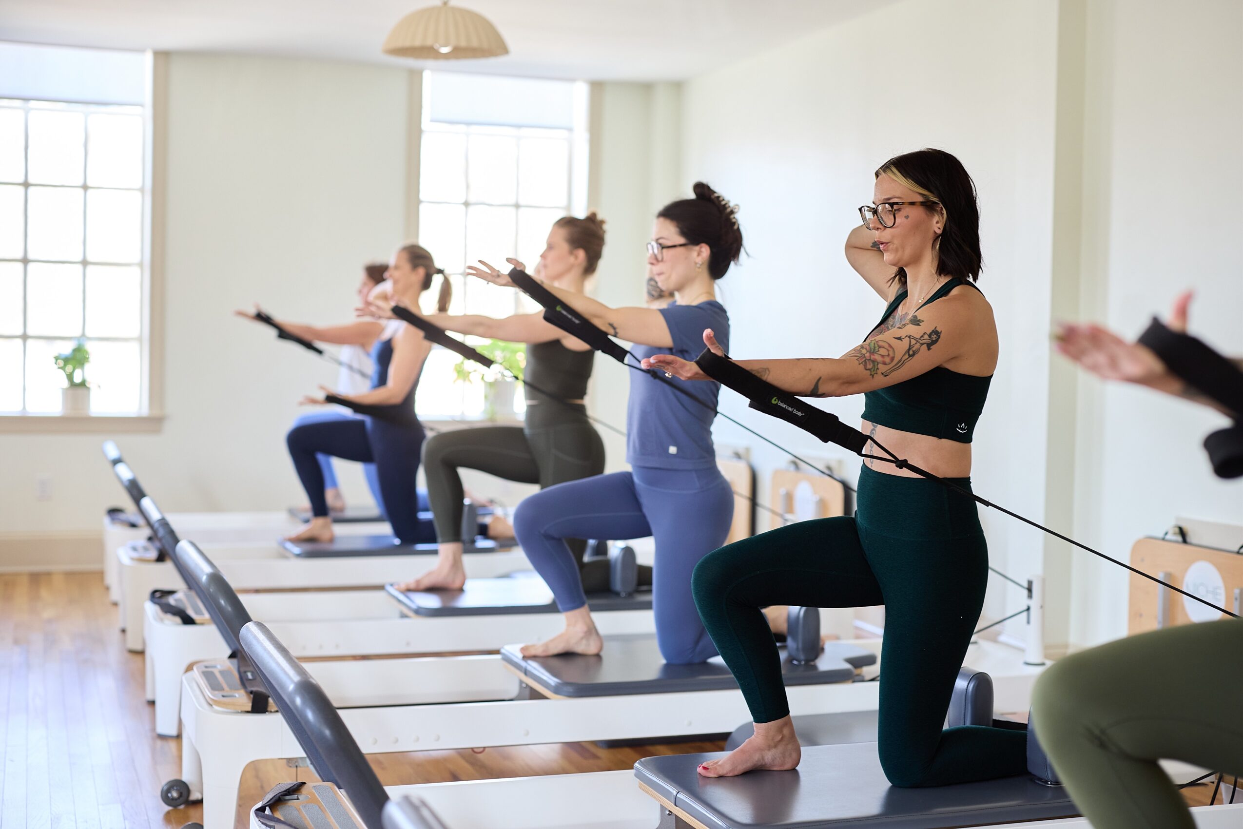 Group of women practicing Pilates on reformer machines in a bright studio with large windows.