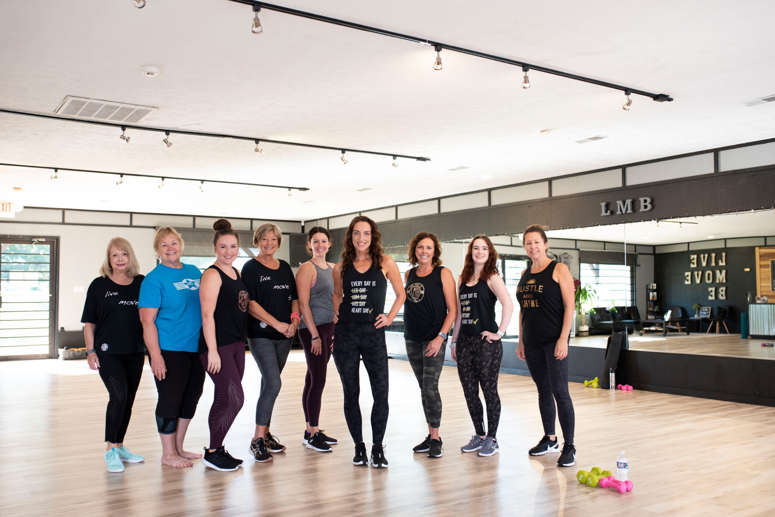 Group of nine women standing in a dance studio, smiling, with a stage and large windows in the background.