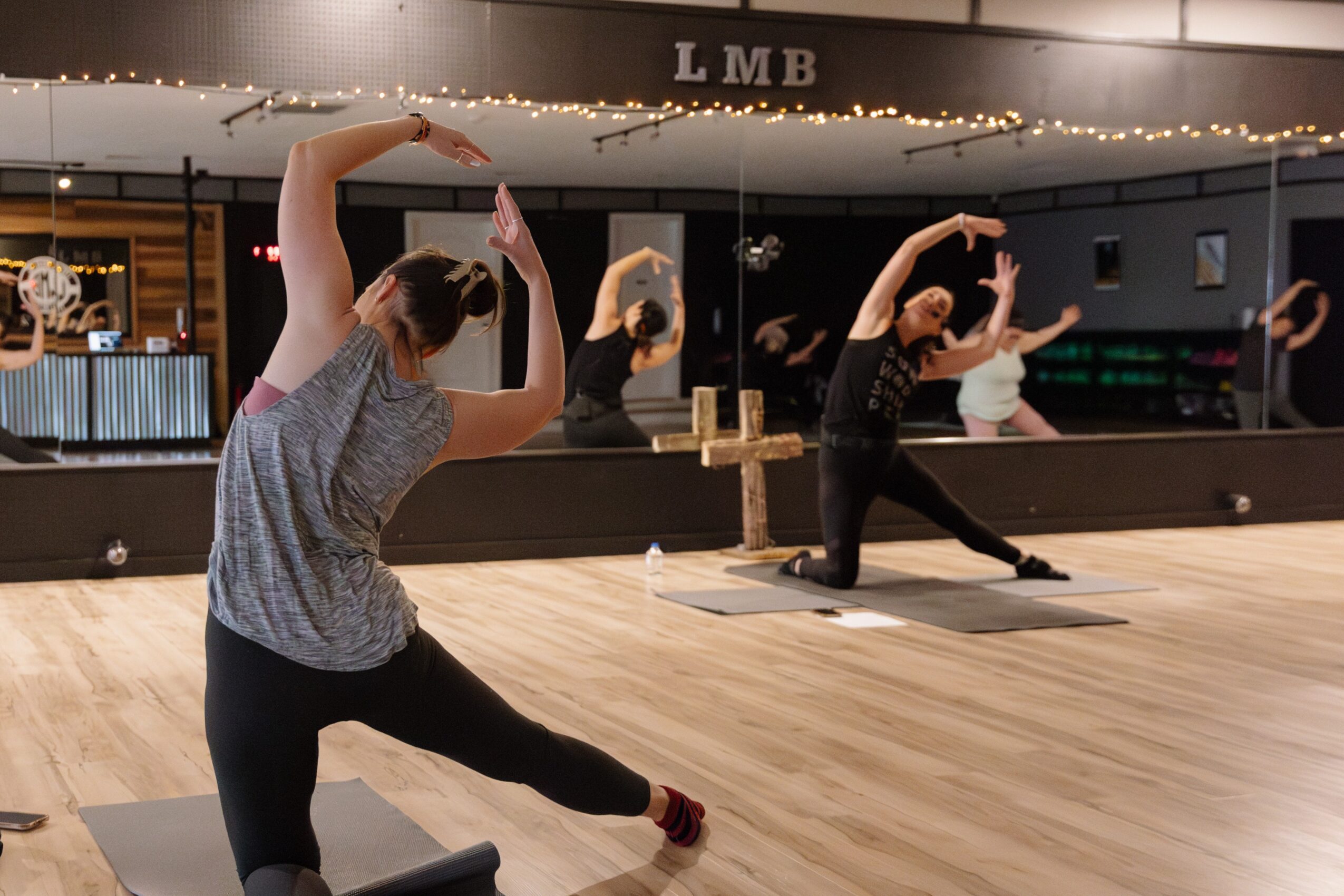 Four people practicing yoga in a dance studio with a mirror, wooden floor, and string lights, stretching with arms raised overhead.