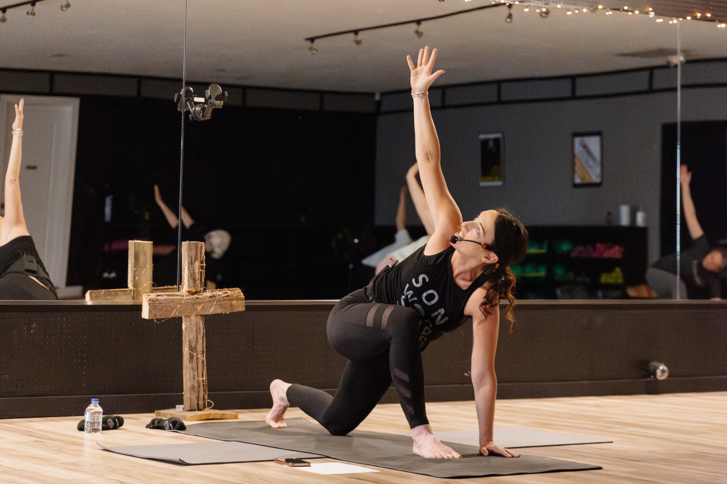 Person practicing yoga or stretching in a studio with mirrors, wearing black workout clothes, raising one arm upward.