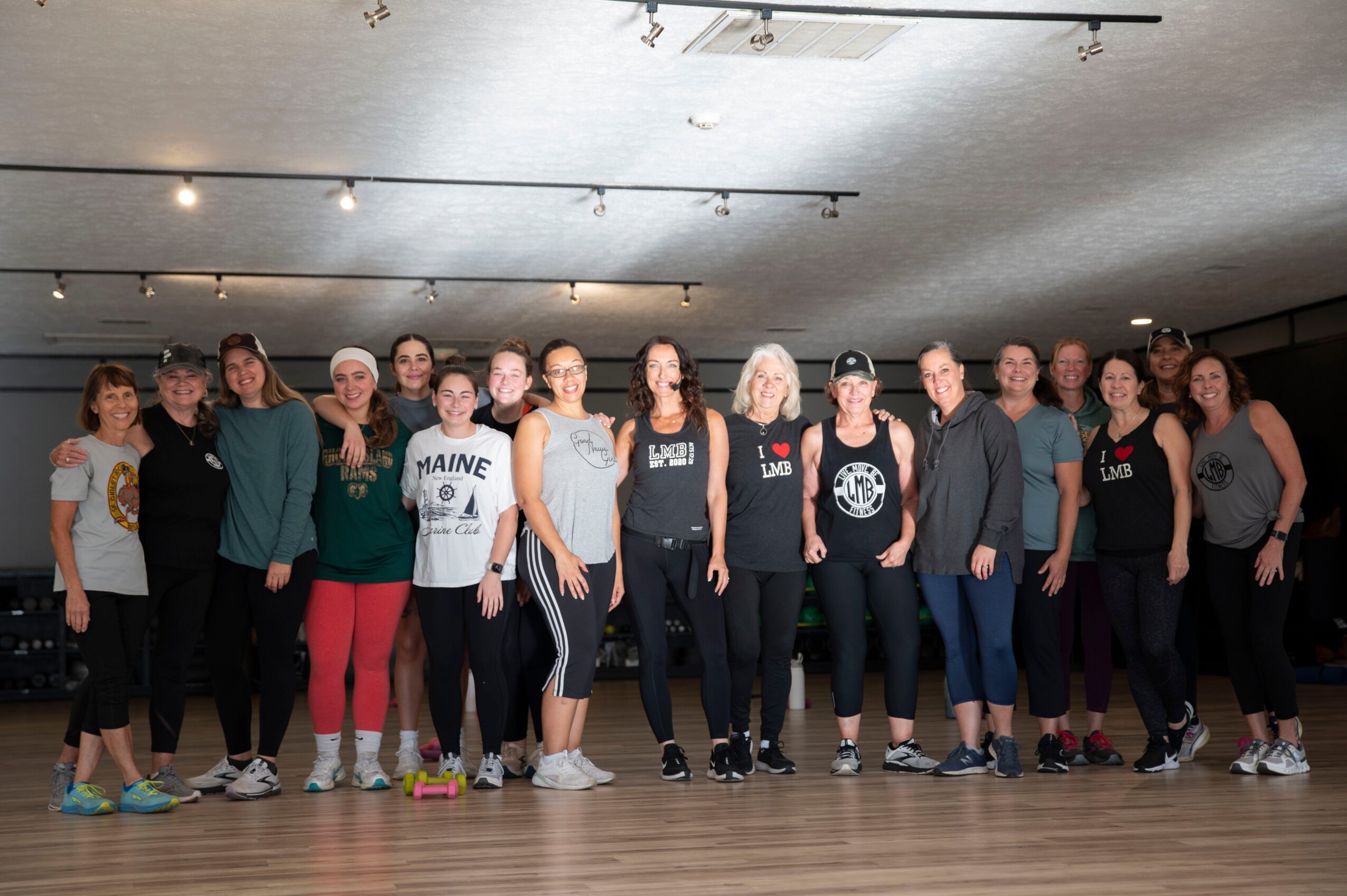 Group of people standing in a dance studio, some with hands on hips, smiling, with a wooden floor and ceiling lights.
