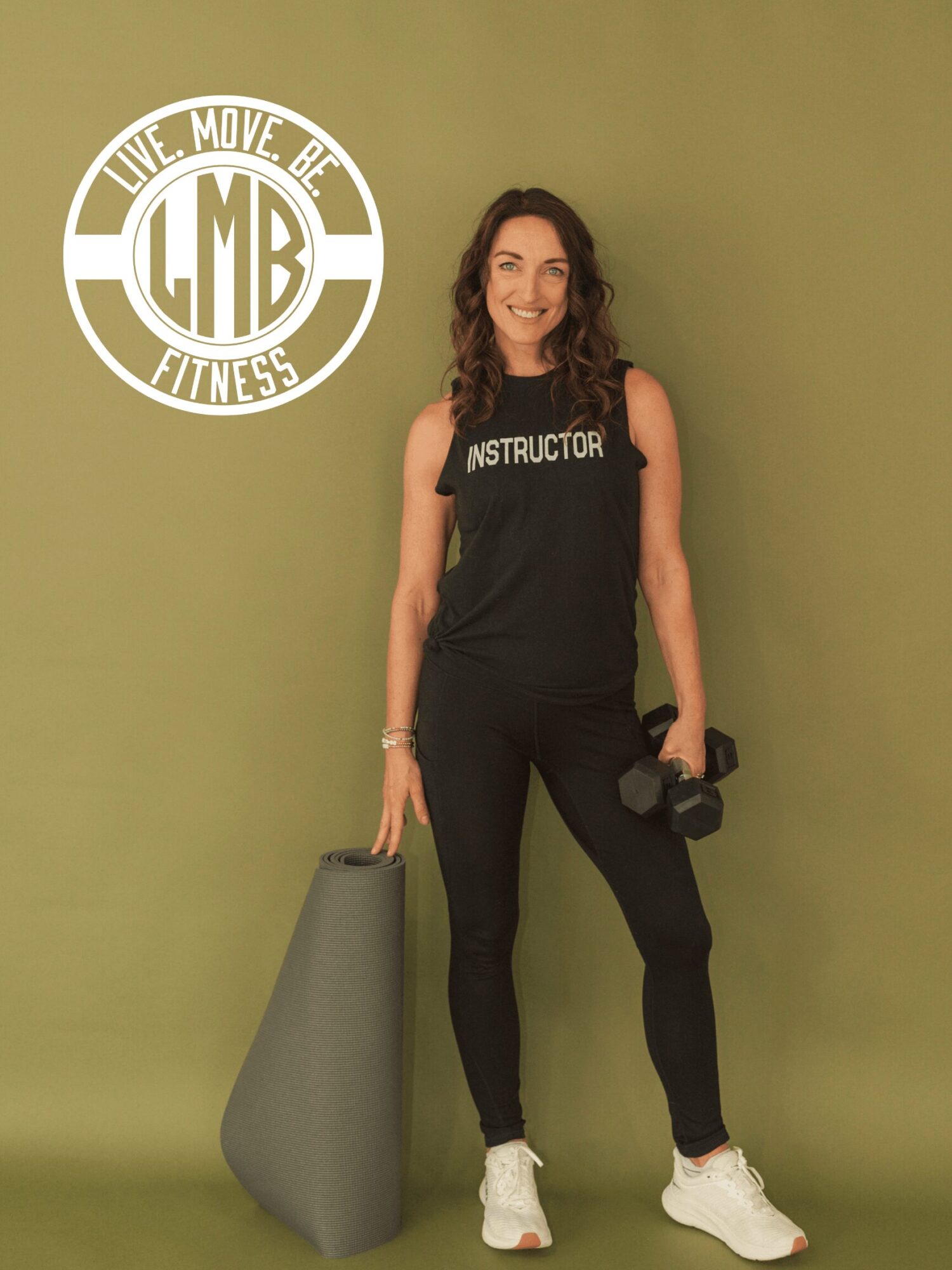 Woman in fitness instructor attire holding a dumbbell, standing next to a foam roller, smiling, against a green wall.