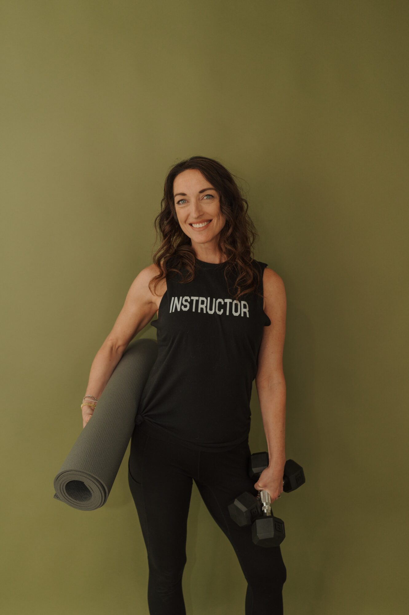 Woman smiling holding a yoga mat and a dumbbell, wearing a black tank top with 'INSTRUCTOR' text, standing against a green background.