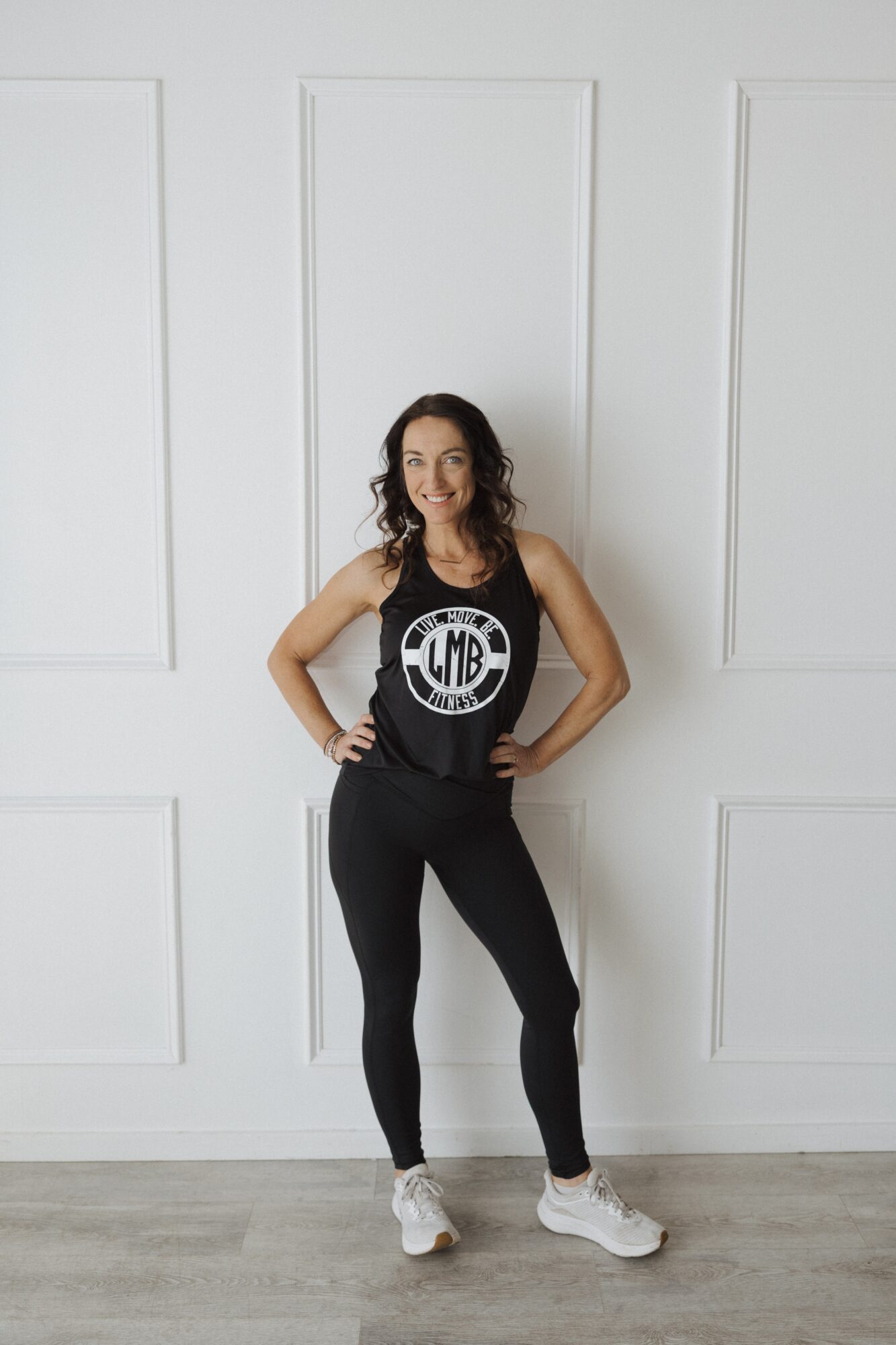 Woman standing with hands on hips, smiling, wearing a black tank top and leggings, in front of a white wall.