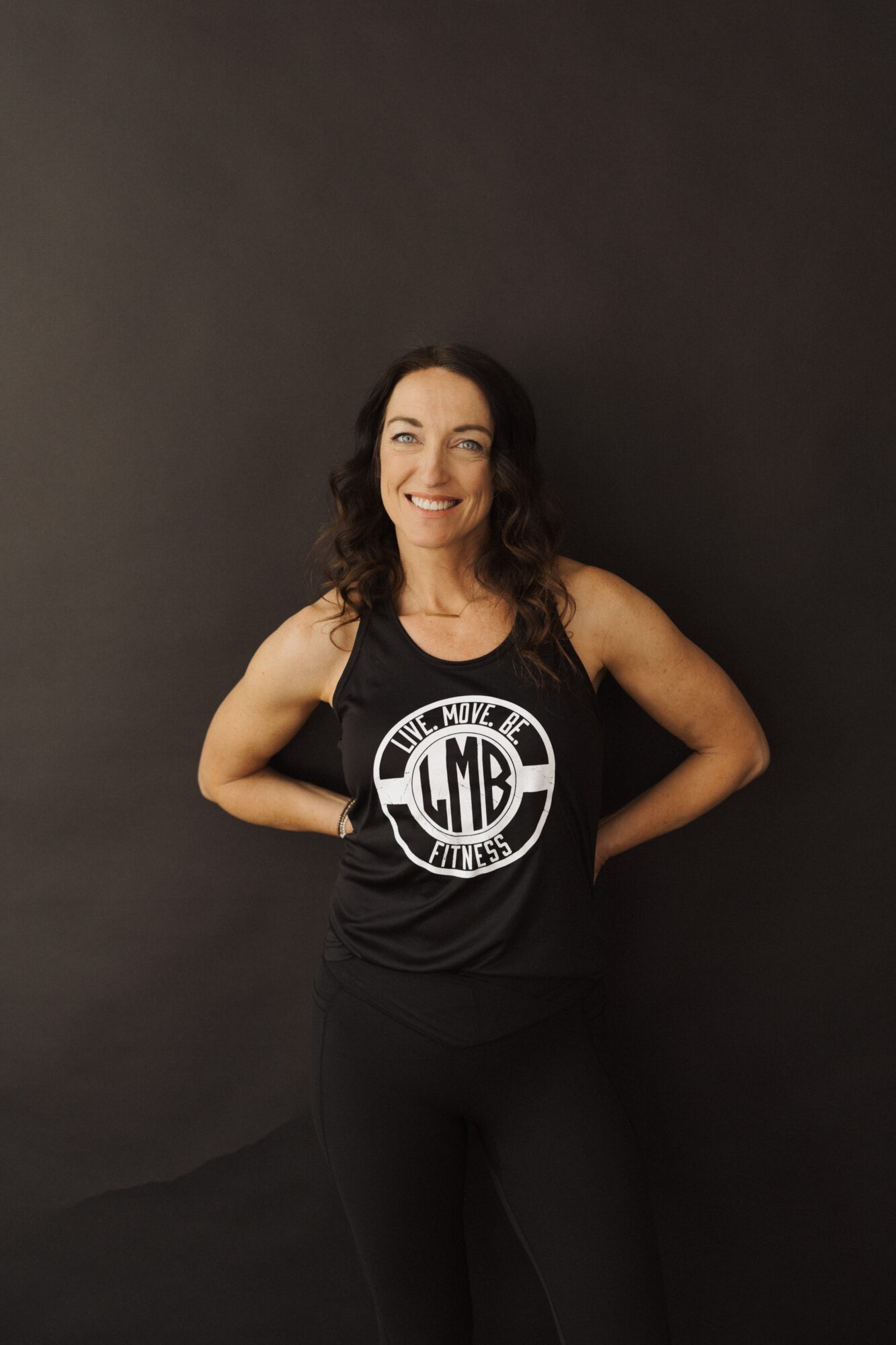 Woman smiling, standing with hands on hips, wearing a black tank top with a logo, against a dark background.