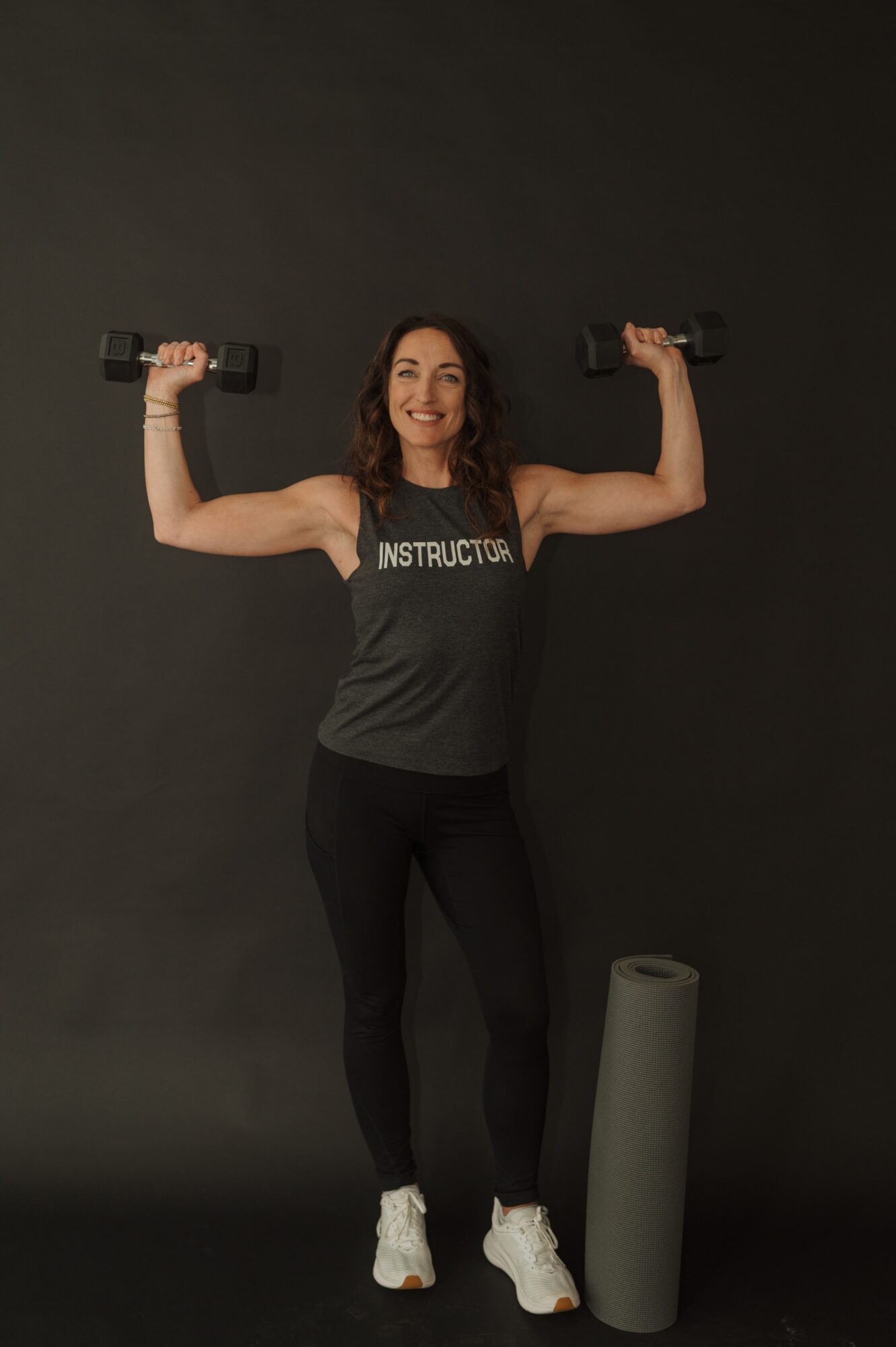 Woman holding dumbbells with arms raised, smiling, standing next to a foam roller against a dark background.