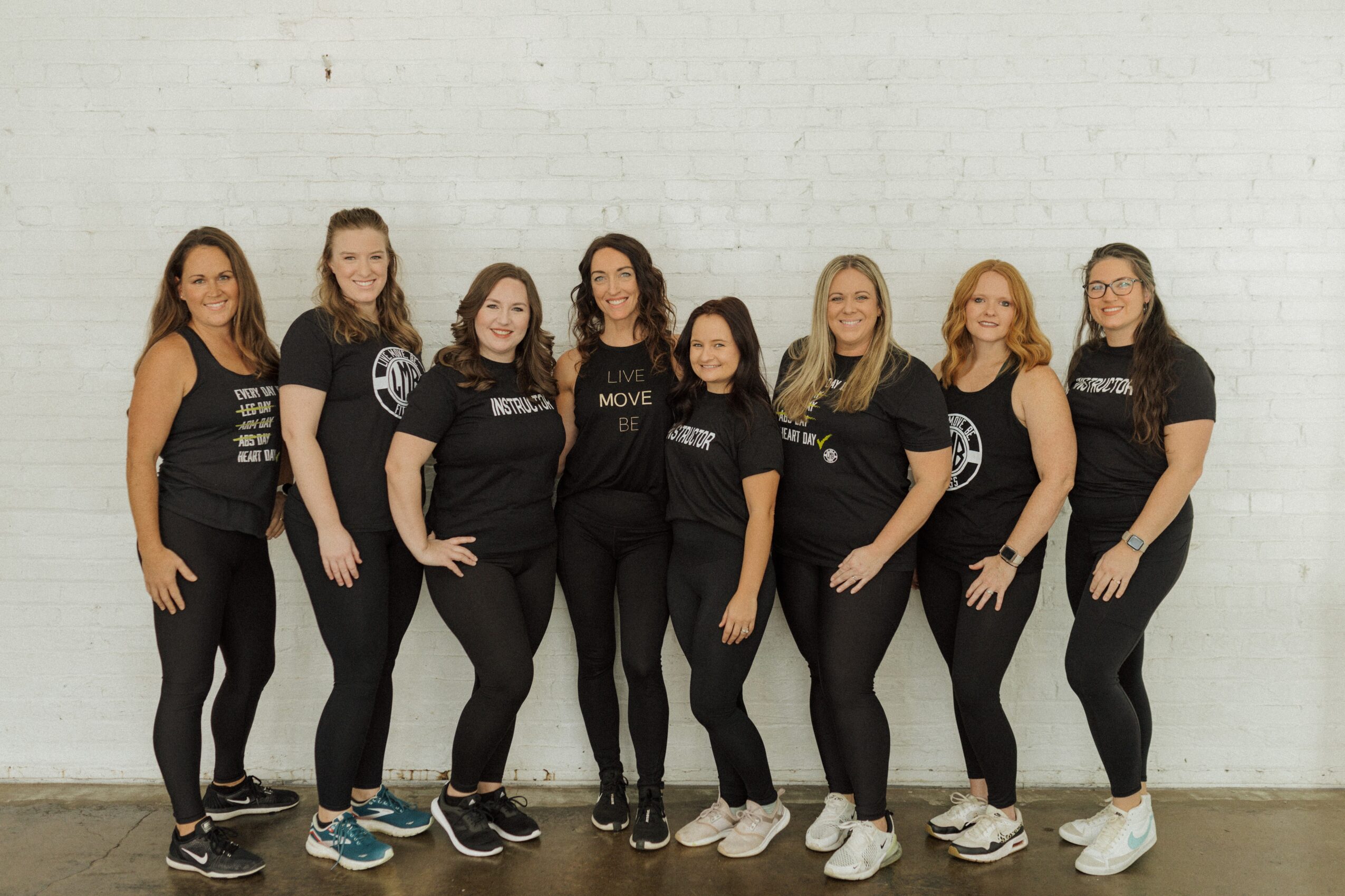 Seven women stand in a line against a white brick wall, smiling and wearing black t-shirts and pants.
