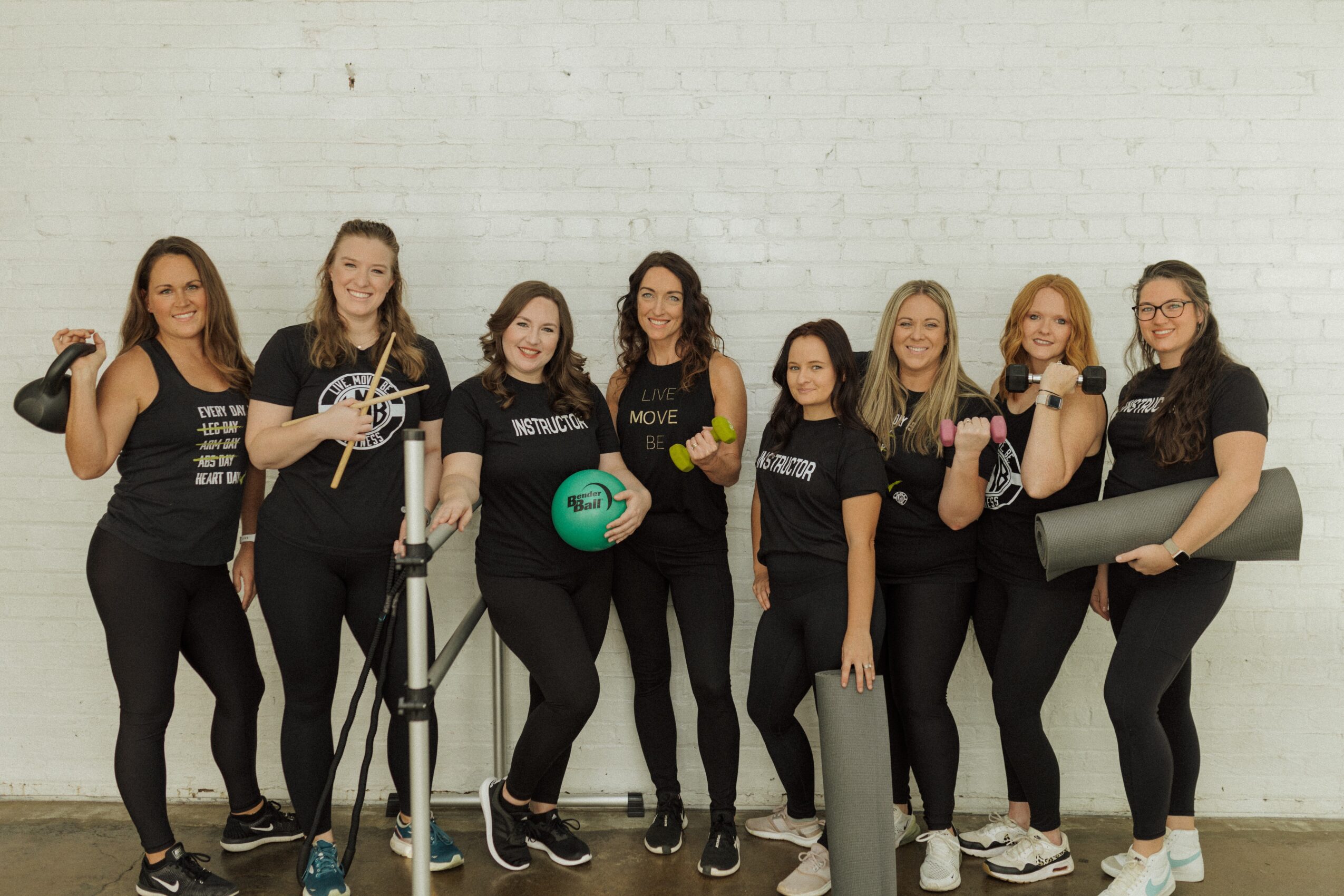 Group of seven women in black workout clothes standing against a white brick wall, some holding fitness equipment.