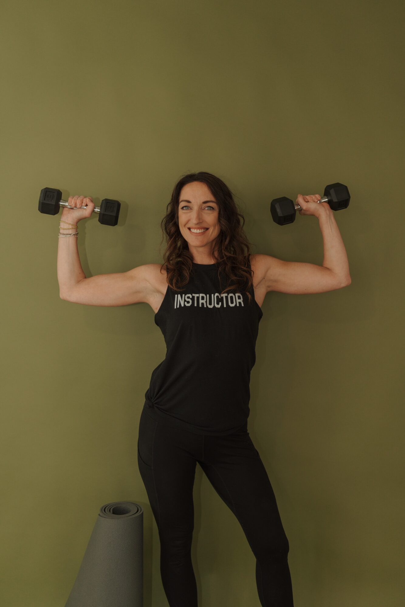 Woman smiling, holding dumbbells at shoulder height, wearing a black tank top with 'INSTRUCTOR' text, standing against a green wall.