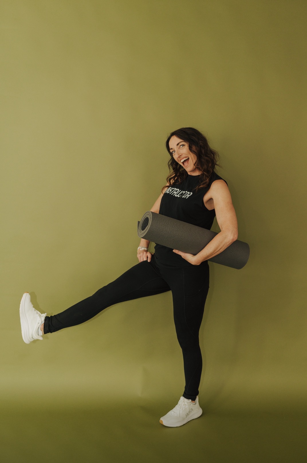 Woman standing against a green wall, holding a yoga mat, with one leg extended sideways, smiling.