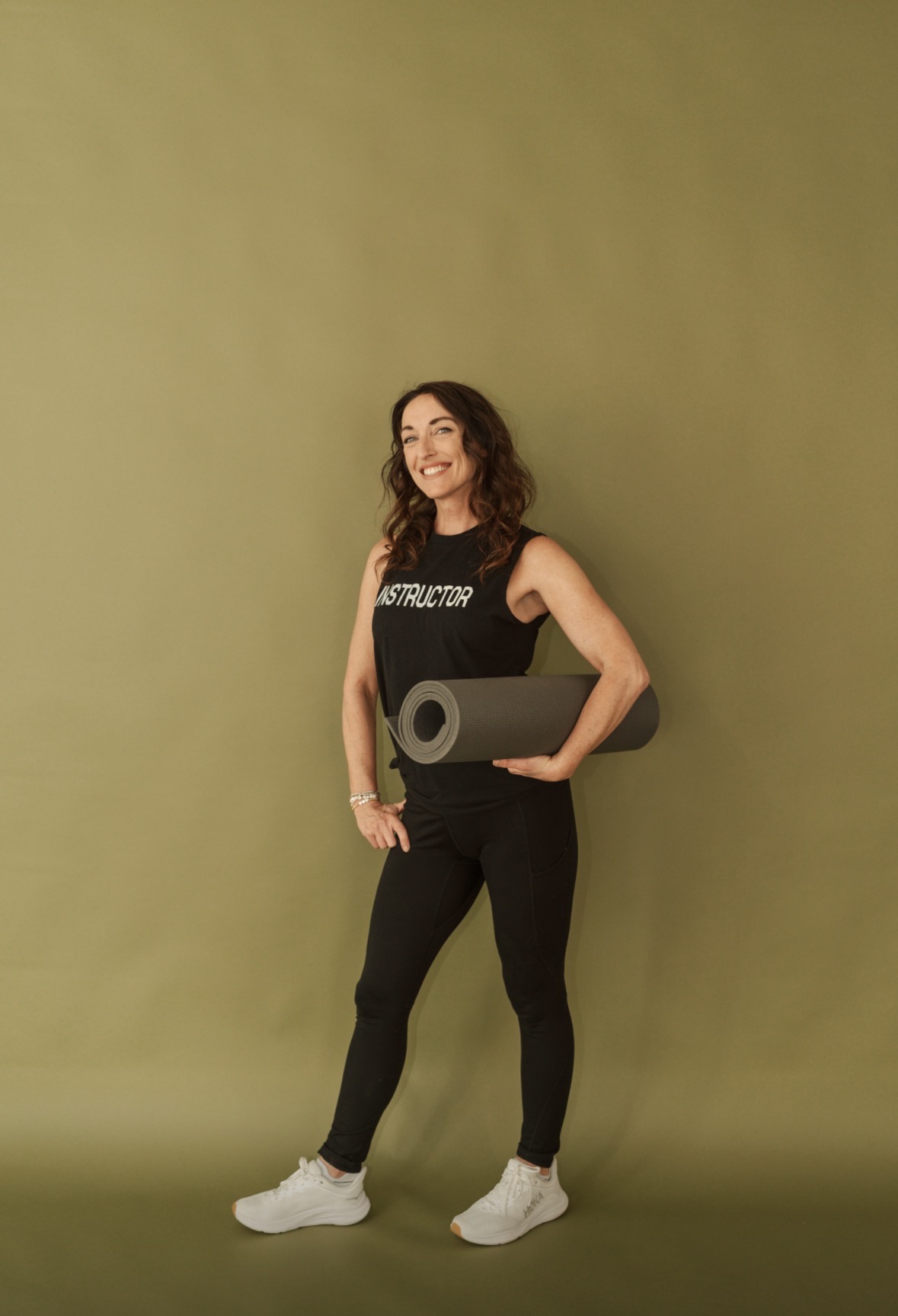 Woman standing against a plain green background, holding a yoga mat, smiling, wearing a black sleeveless top and black pants.