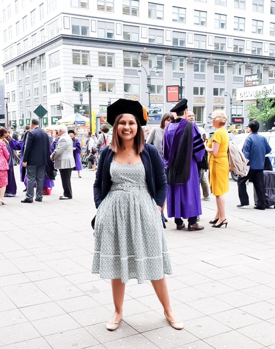 Young woman in a dress and graduation cap smiling in a city square with people and buildings in background.