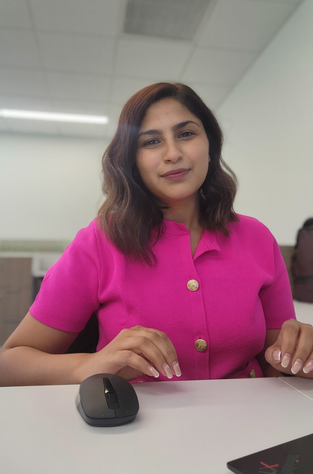 Young woman with shoulder-length wavy hair wearing a bright pink top, sitting at a desk with a computer mouse, in an indoor setting.