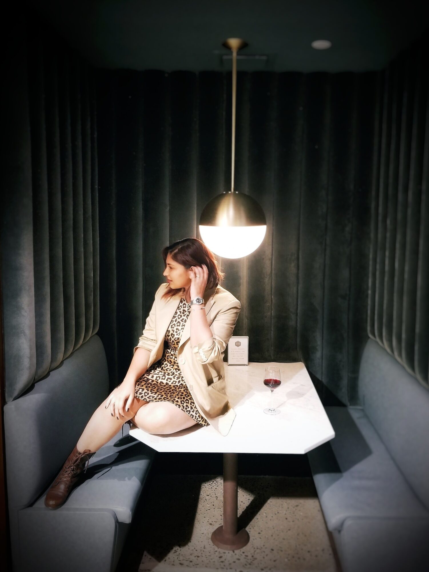 Woman sitting at a table in a dark, enclosed space with curtains, illuminated by a hanging light, wearing a beige blazer and leopard print dress.