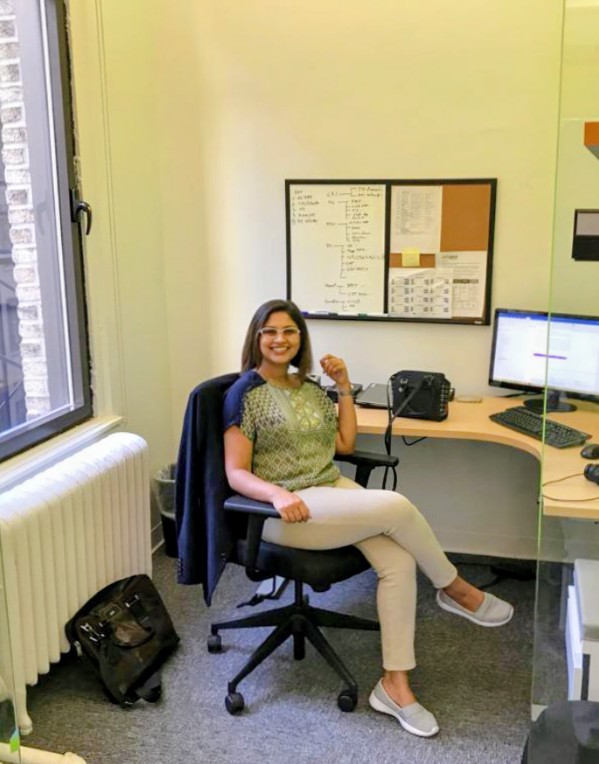 Woman sitting at desk in office, smiling, with computer monitors and a corkboard behind her.
