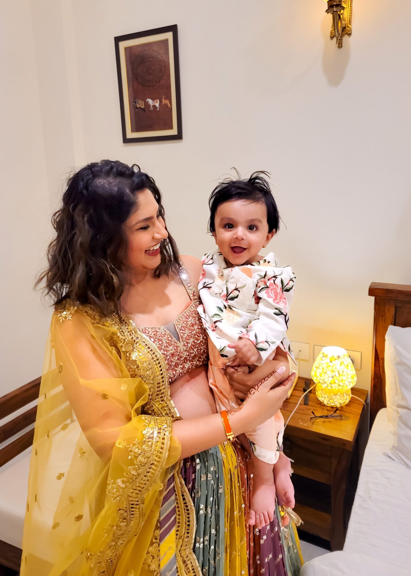 Woman holding a smiling baby girl in a decorated room with a framed picture and a lamp on a side table.