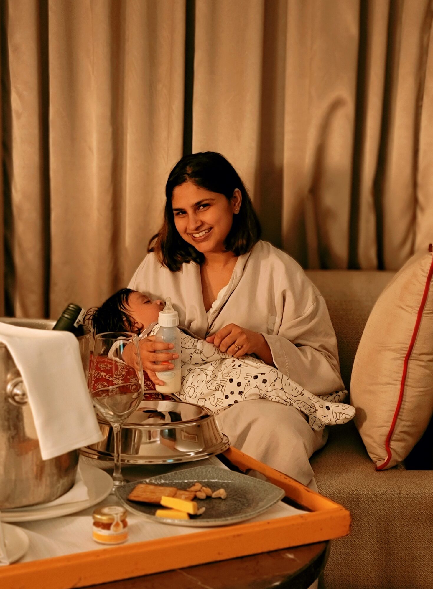 Woman sitting on a sofa, smiling, with a tray of food and drinks in front of her, beige curtains in background.