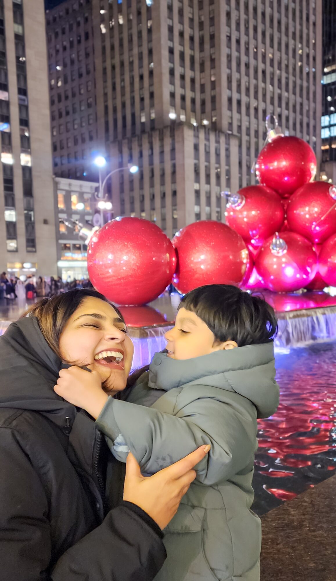 Woman and child smiling and playing near a fountain with large red ornaments and city buildings in the background.