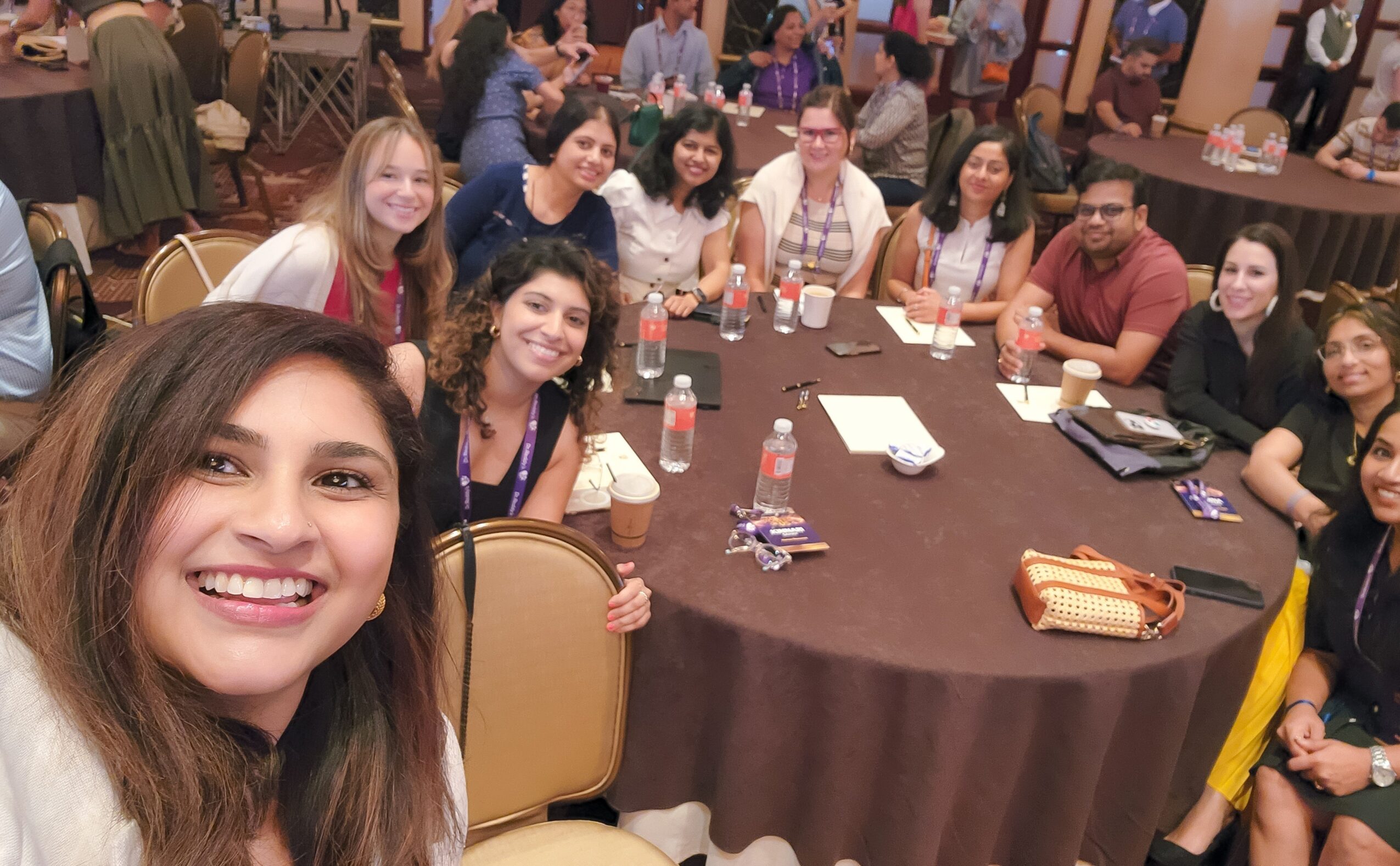 Group of people gathered around a round table at a social event, smiling and taking a selfie.