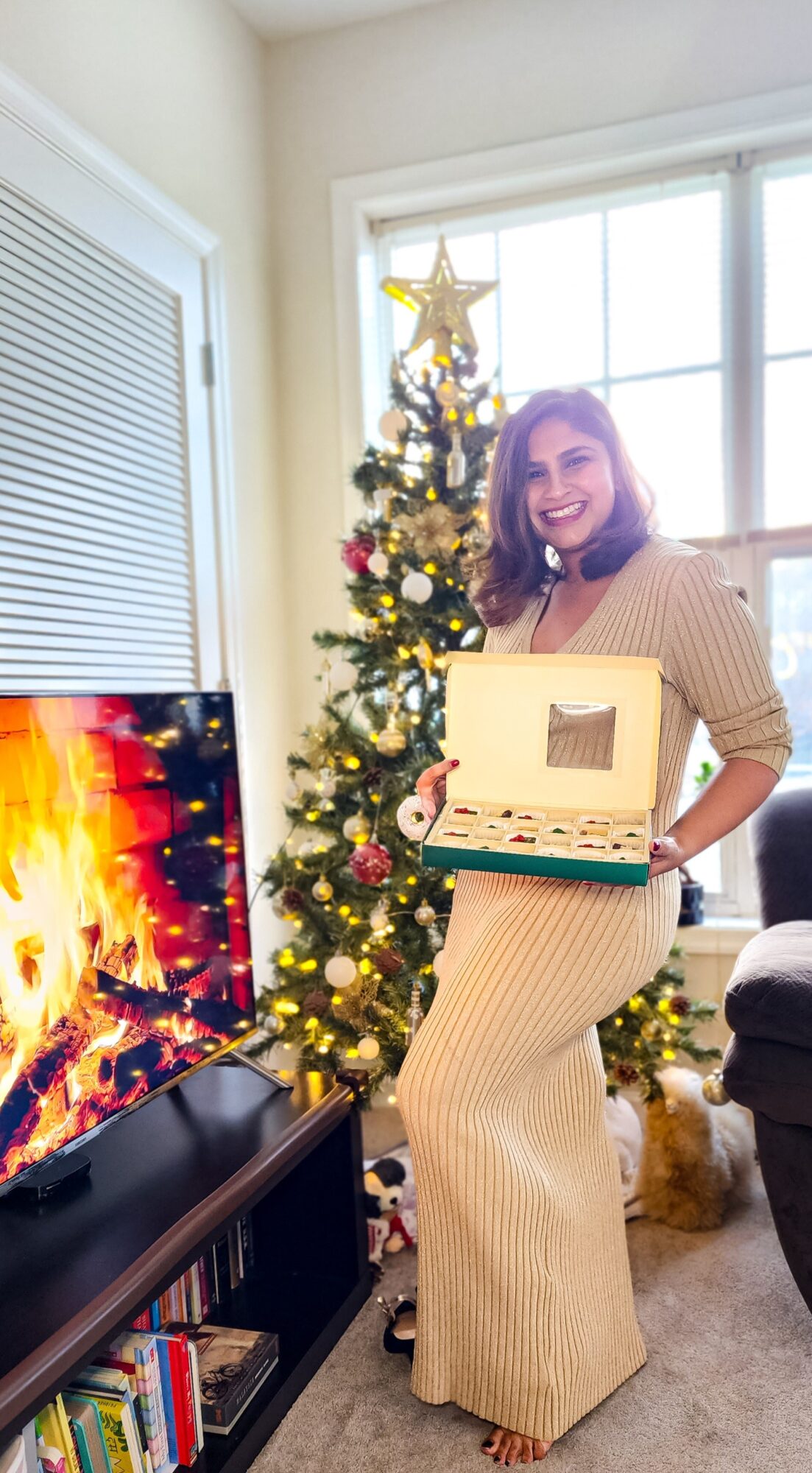 Woman in beige dress holding a box, standing next to a decorated Christmas tree, smiling, in a living room with a fireplace and TV.