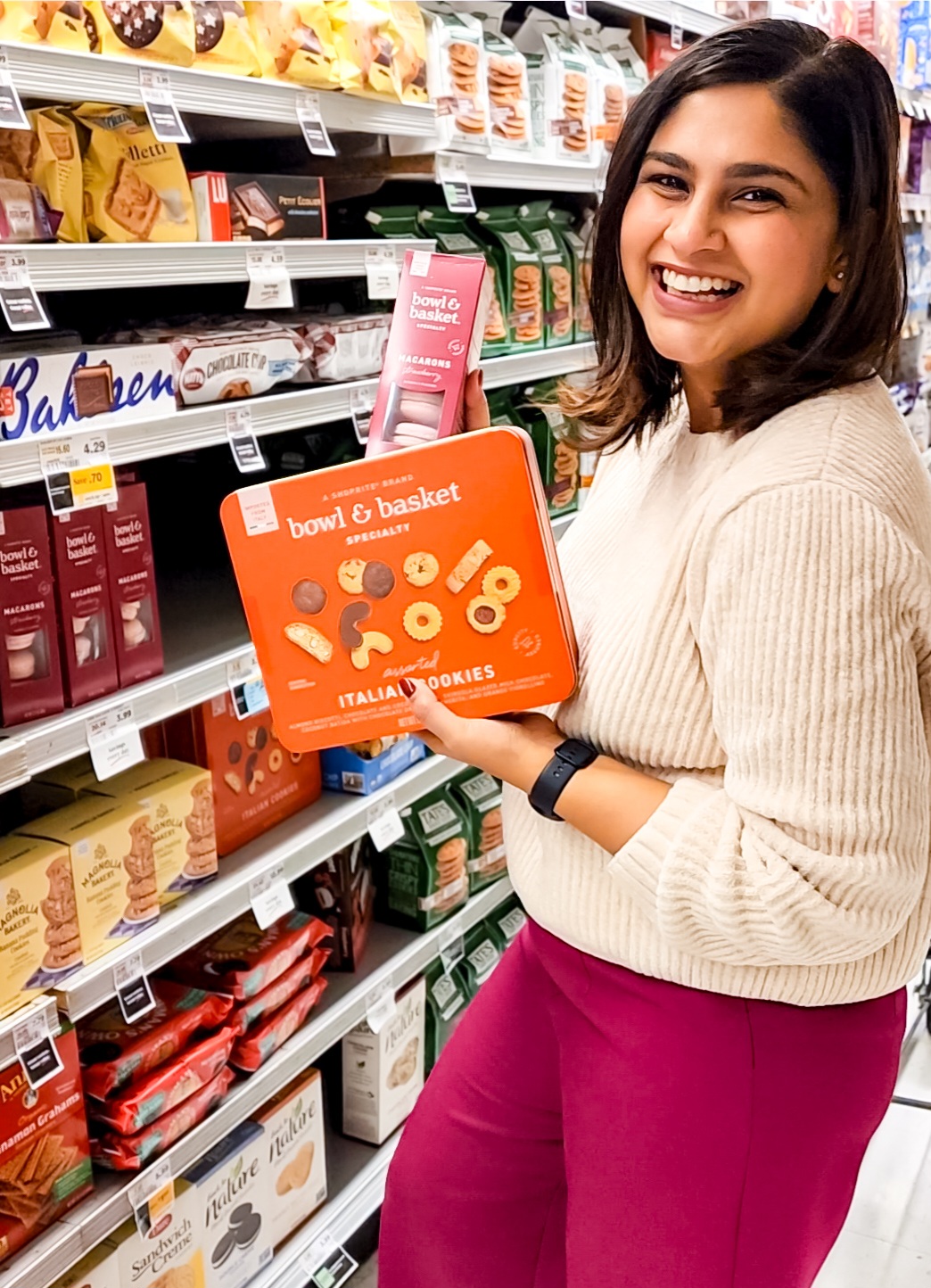 Smiling woman holding a boxed product in a grocery store aisle with shelves of food items.