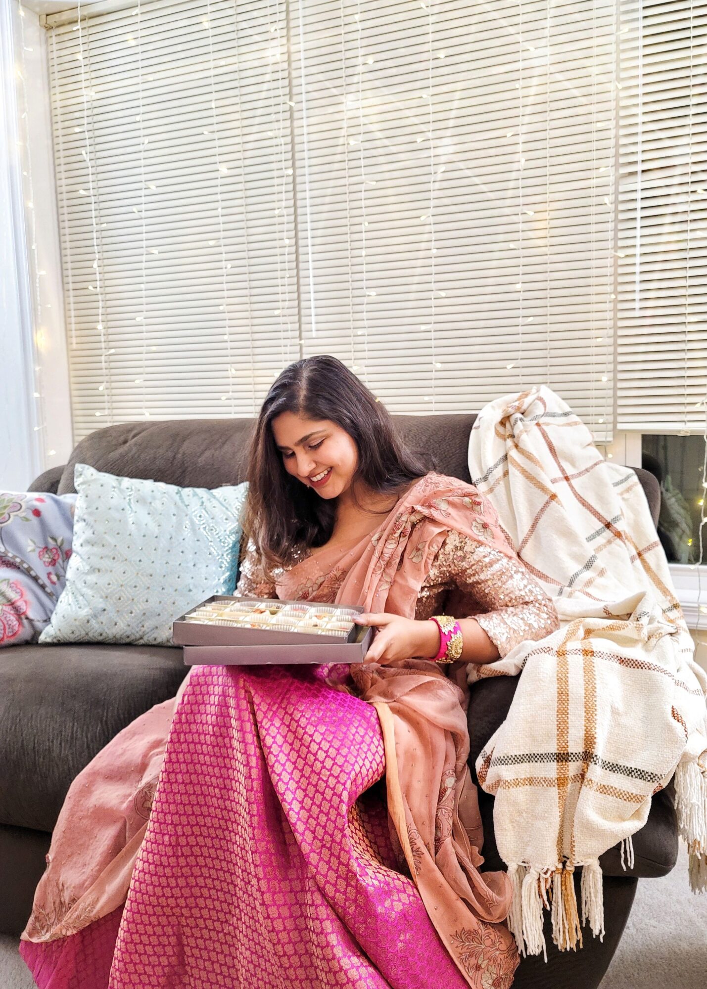 Young woman sitting on a sofa, reading a book, with pillows and a blanket nearby, in a bright room with blinds.