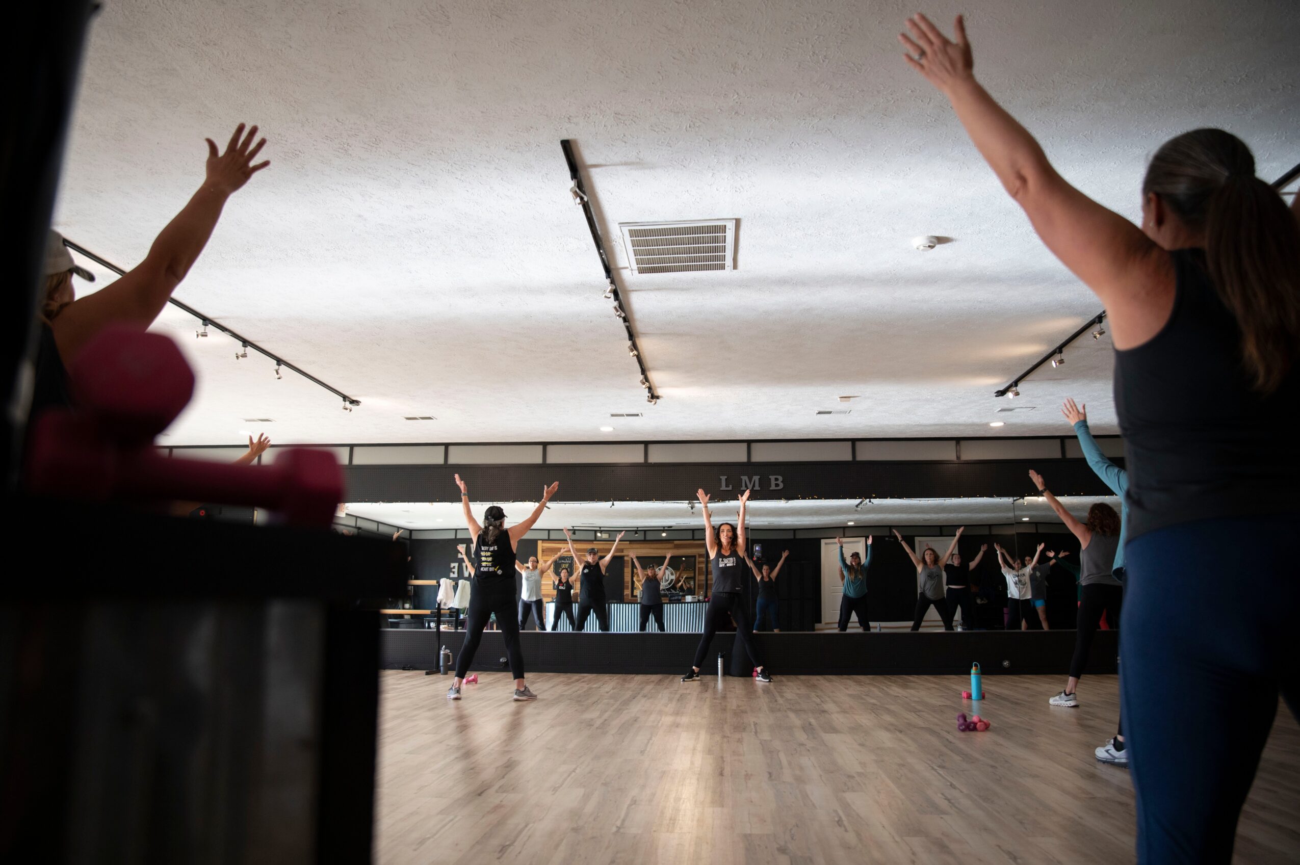 People participating in a fitness class with arms raised, in a room with a large mirror and wooden floor.