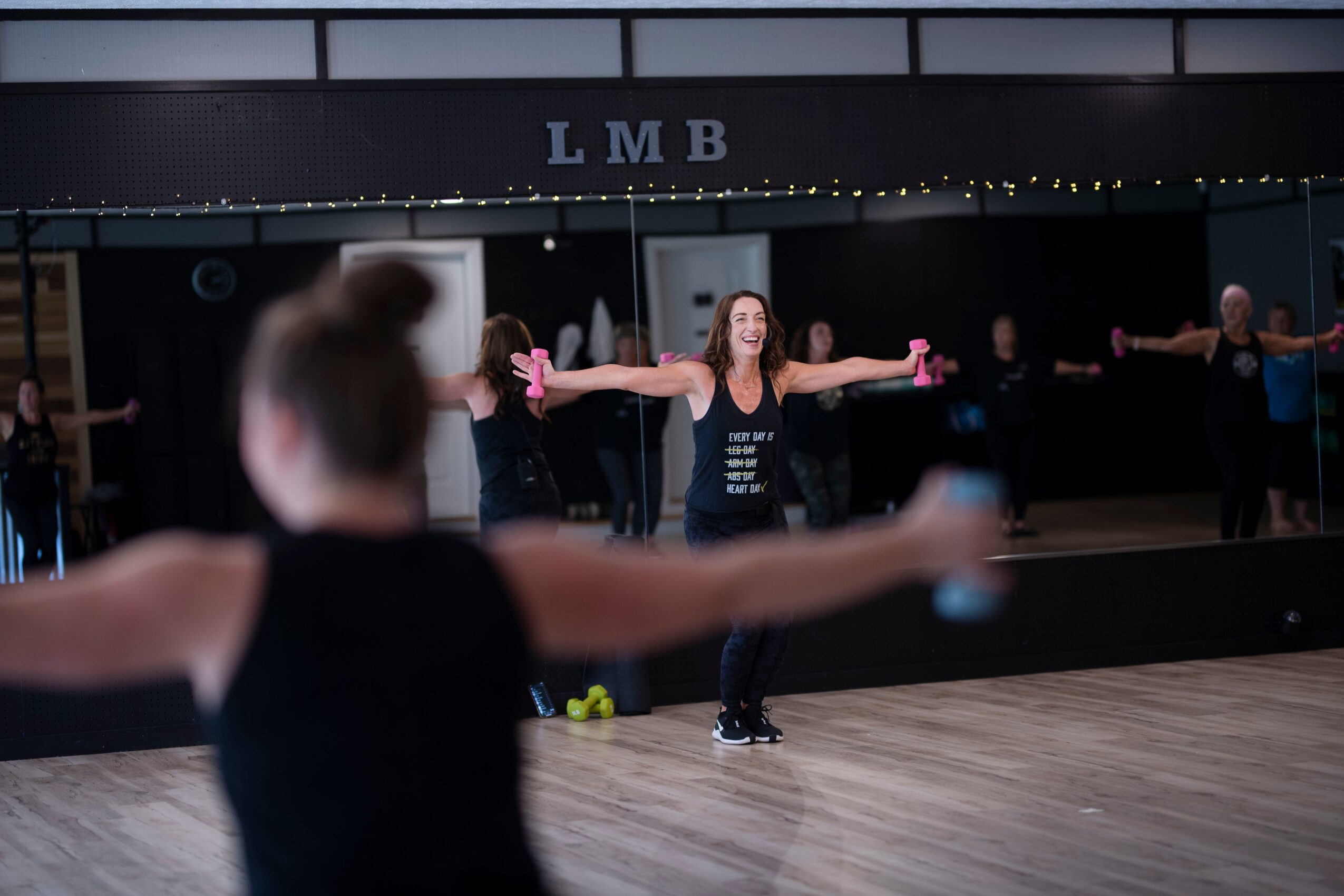 People in a dance studio with a mirror, woman smiling with arms outstretched, others in the background, wooden floor.