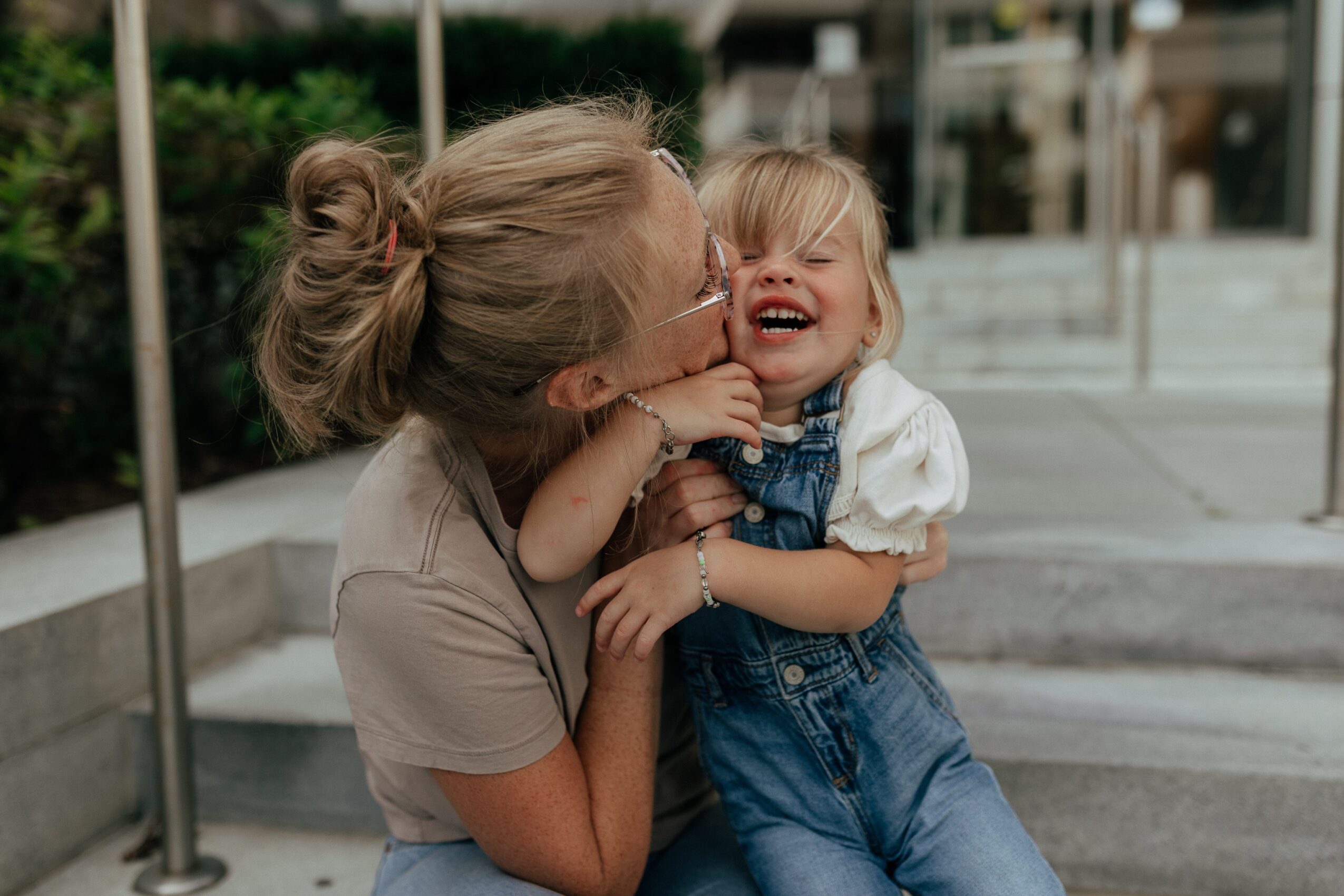 Adult woman and young girl laughing and hugging outdoors on a sidewalk, with greenery and buildings in the background.