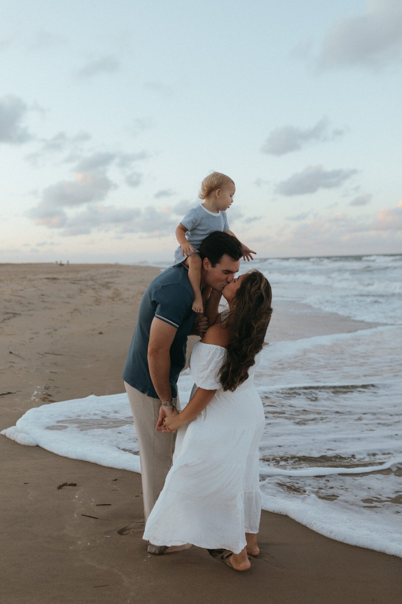Family on the beach, a man and woman holding hands, with a child sitting on the man's shoulders, ocean waves in background