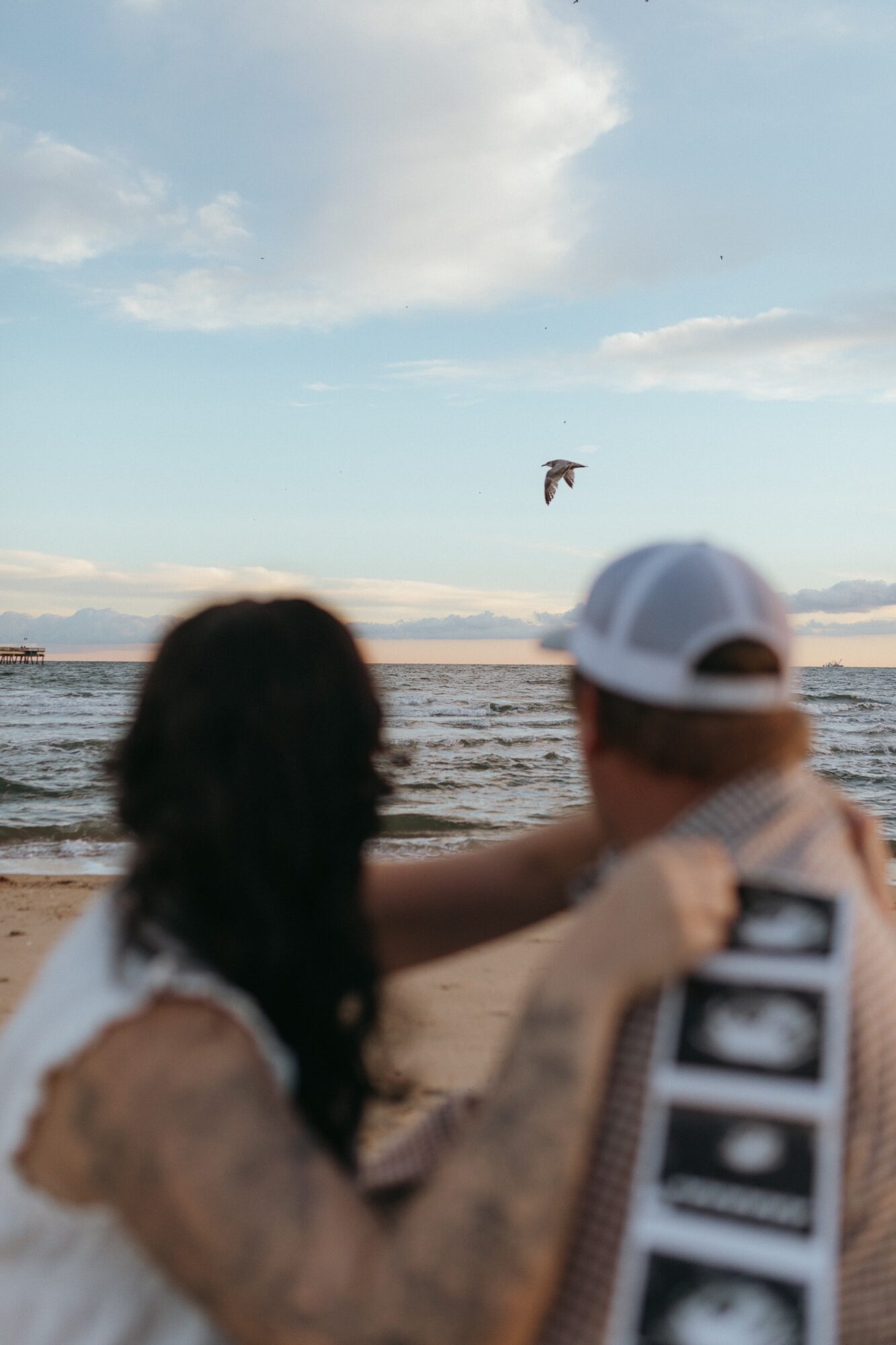 Two people on a beach, one with long dark hair and the other wearing a white cap, watch a bird flying overhead.