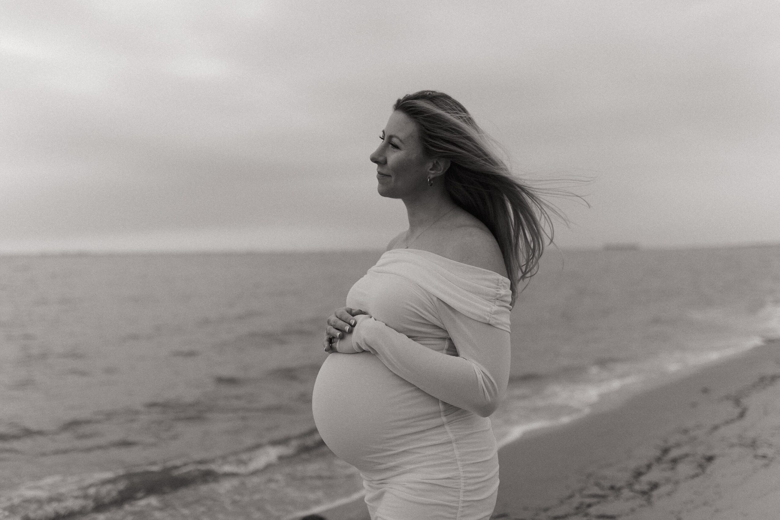 Pregnant woman standing on a beach, looking to the side with wind blowing her hair, in black and white.