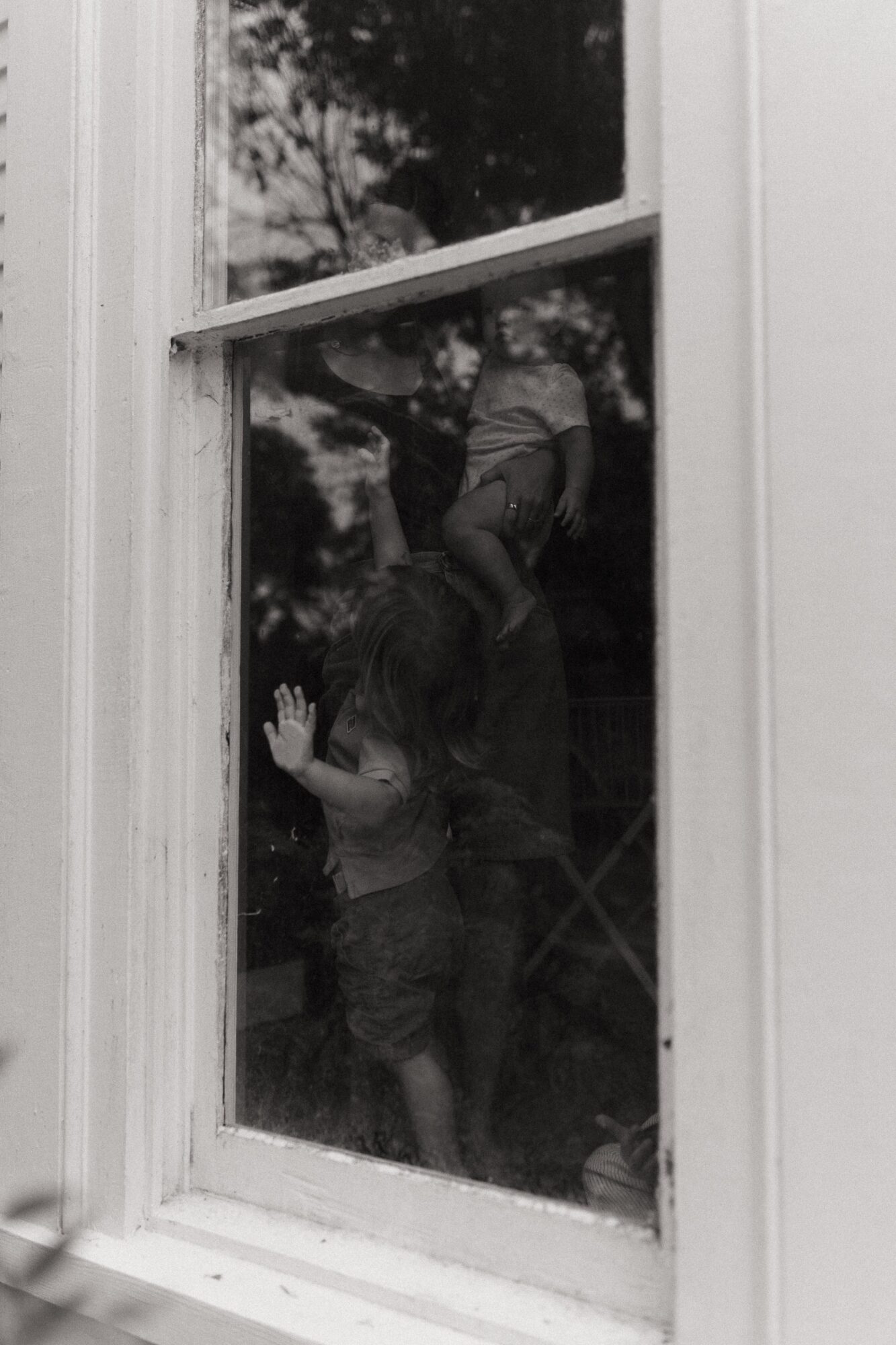 Two children looking out a window, one with hand on glass, the other sitting on a ledge outside, trees visible through the window.
